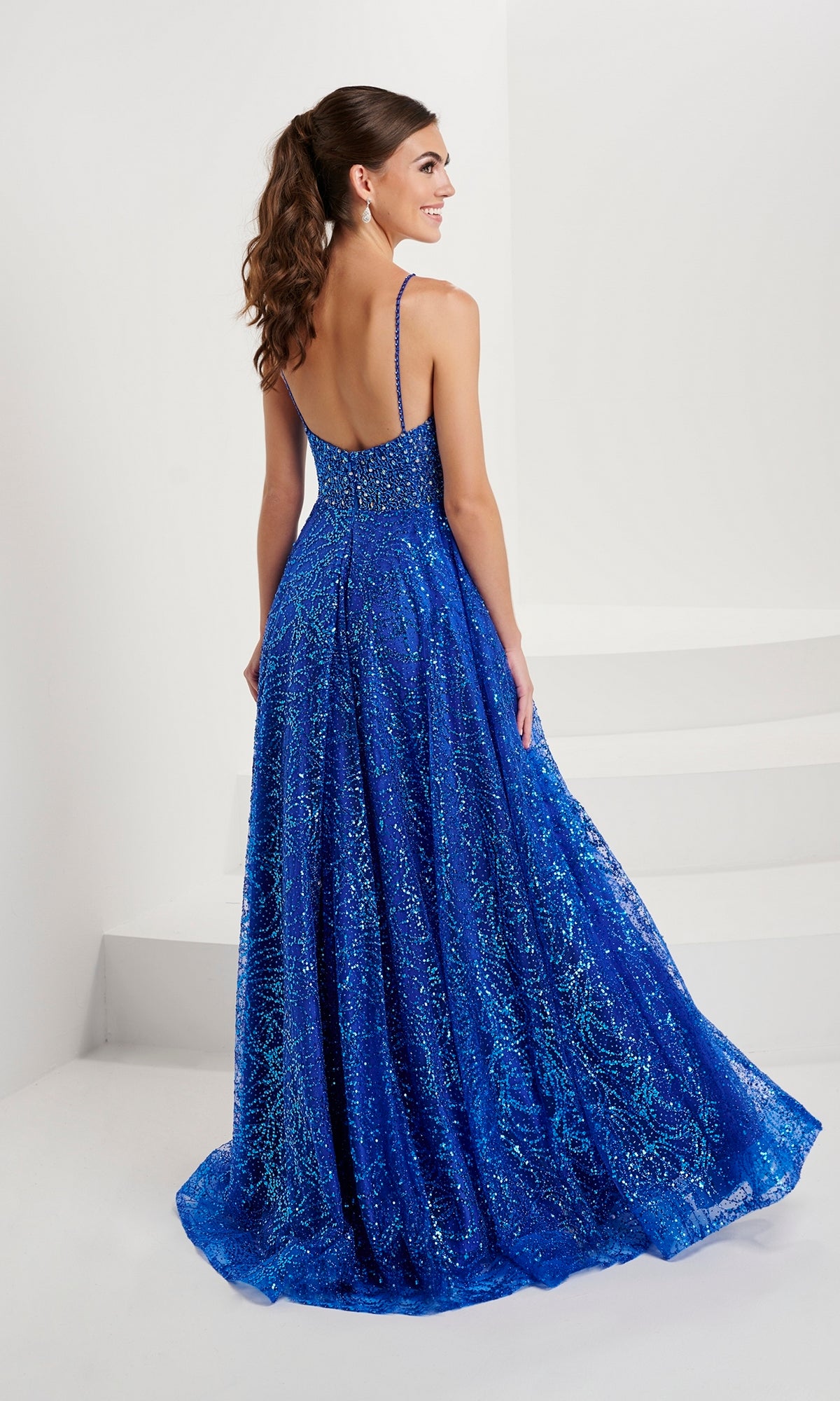 A woman with long brown hair in a ponytail wears the Tiffany Beaded-Bodice Glitter Tulle Prom Dress 16088, featuring an A-line silhouette and thin straps, as she stands on a white floor near white stairs.