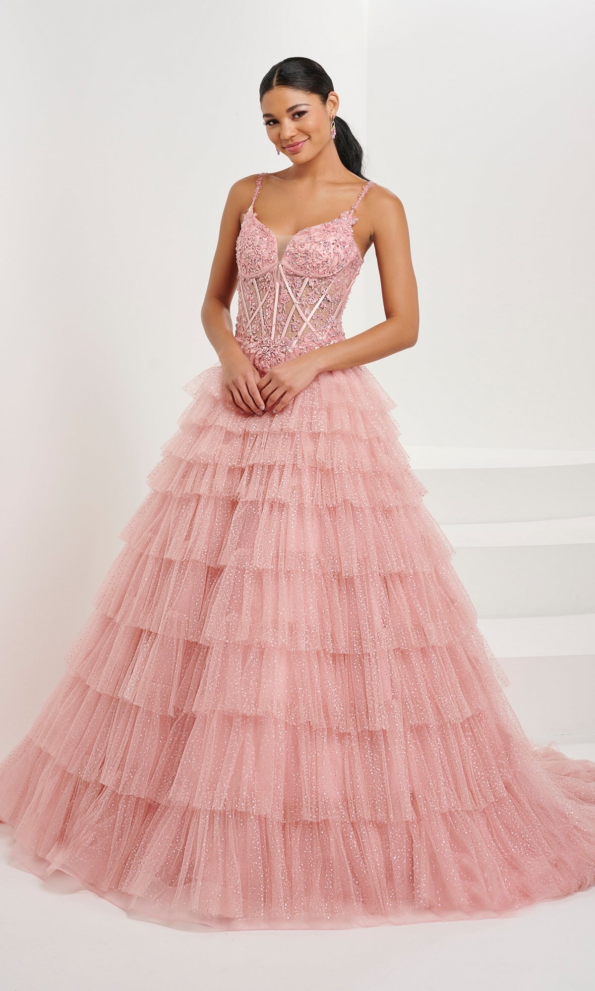 A woman stands indoors wearing the Tiffany Ruffled Glitter Tulle Prom Ball Gown 16099, featuring a pink beaded corset bodice with thin straps and a tiered glitter tulle skirt for extra elegance.