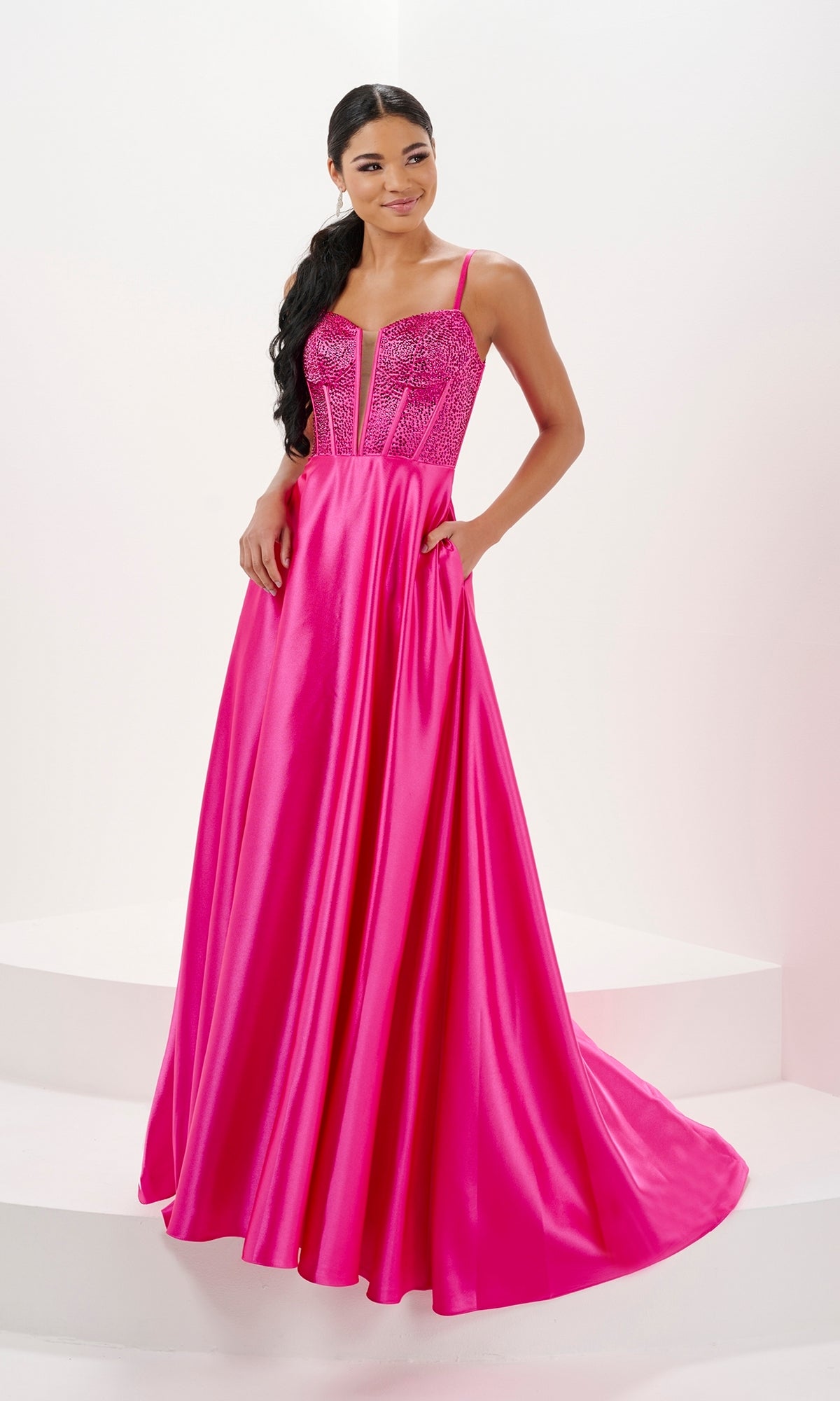 A woman wears the Tiffany Long Jersey A-Line Corset Prom Dress 16101 in bright pink, featuring a sequined corset bodice and thin straps, standing on a white background.