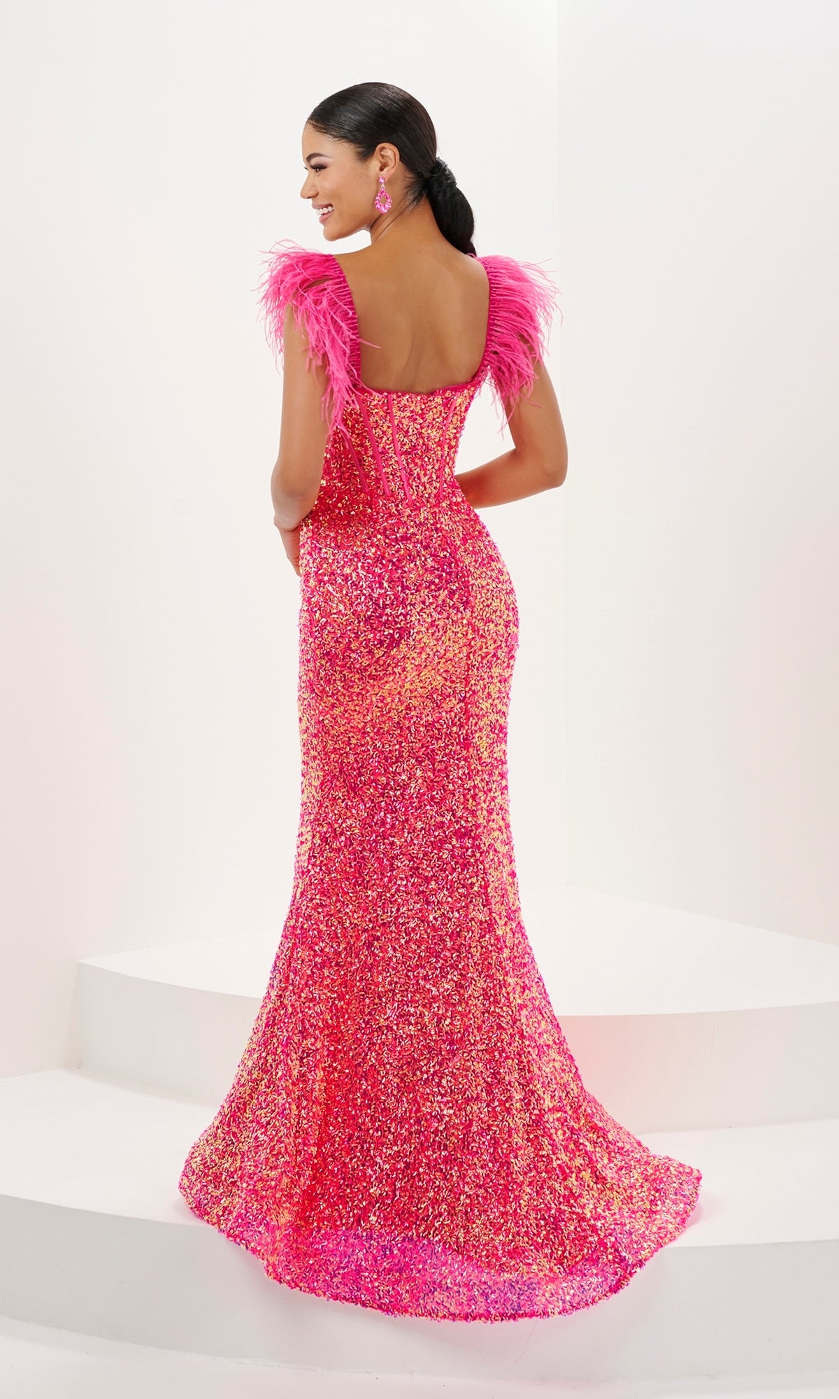 A woman smiles on white steps, facing away in the Tiffany Fitted Sweetheart Sequin Prom Dress 16106—a floor-length, pink, strapless gown with feather straps.