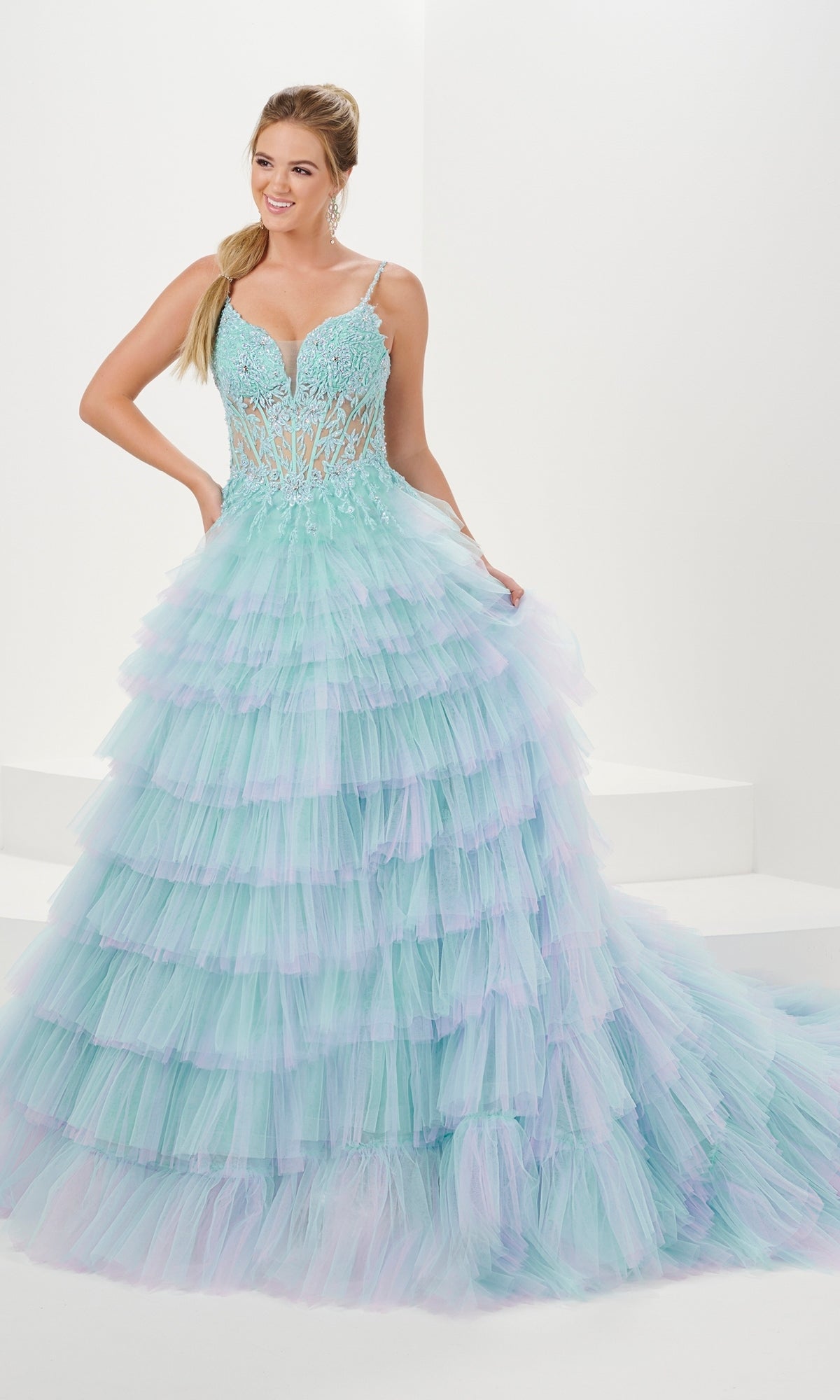 A woman models the Tiffany Tiered Ruffled Tulle Prom Ball Gown 16115, featuring a pastel blue and lavender ombre tulle skirt and embellished sheer corset bodice, standing against a plain white background.