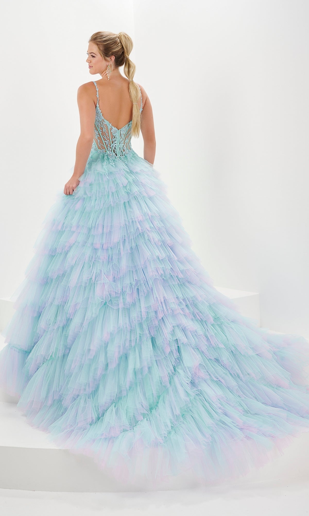 A woman in the Tiffany Tiered Ruffled Tulle Prom Ball Gown 16115, featuring a sheer corset bodice and ombre tulle skirt, stands on white steps with her back to the camera.