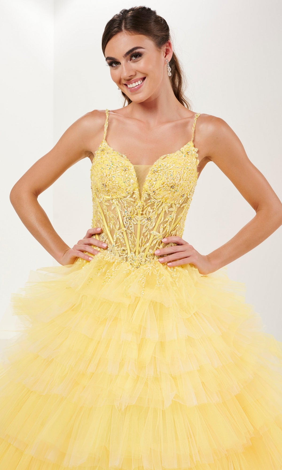 Woman wearing the Tiffany Tiered Ruffled Tulle Prom Ball Gown 16115, featuring a yellow ombre tiered ruffled skirt and floral embroidered bodice, stands with hands on hips and smiles.