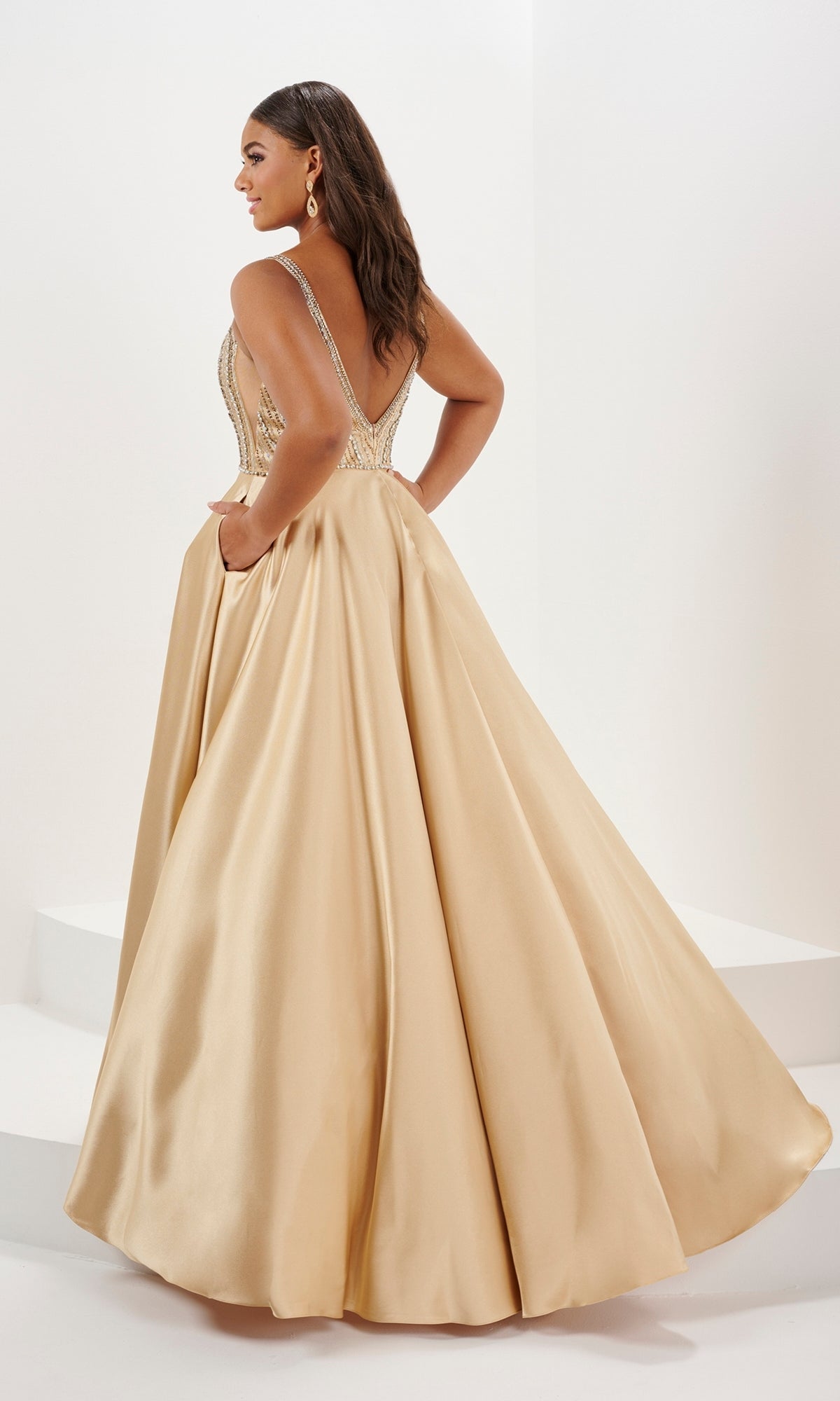A woman models the Tiffany Long Plus-Size Beaded Prom Dress 16129, a sleeveless gold satin A-line gown with beaded straps and a deep V-back, in a minimal white room.