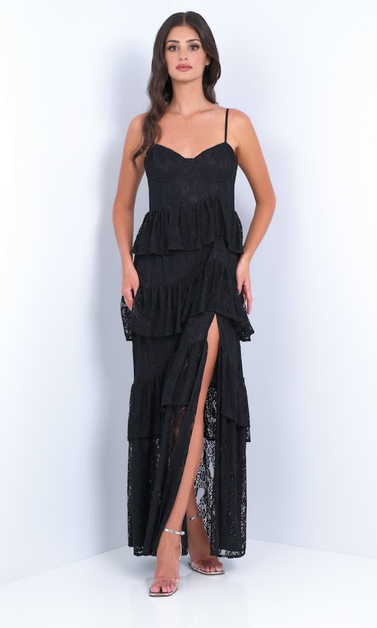 A woman models the Vanna by Velvi: a black ruffled tiered long lace prom dress with spaghetti straps and a front slit, set against a plain white background.