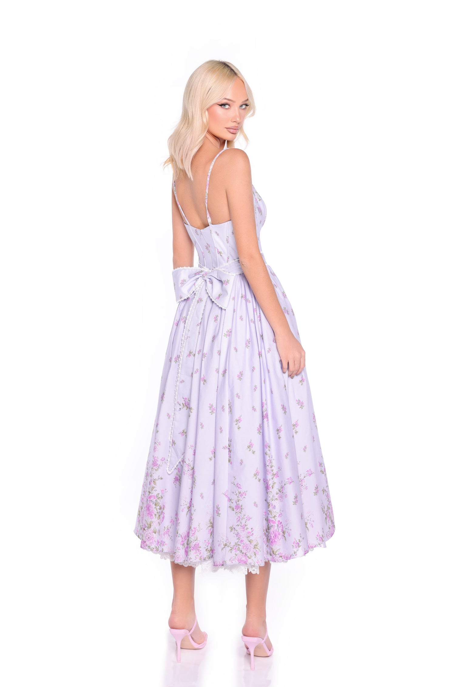 A woman with blonde hair models the Violetta Lilac Purple Midi Casual Dress, which features a corset bodice, floral print, and a large bow at the back. She pairs it with pink heels and looks over her shoulder while facing away.
