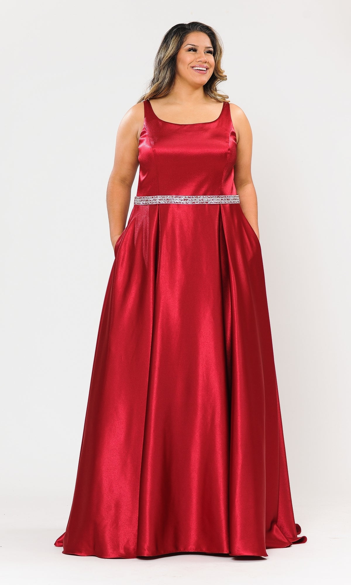 A woman models the Sleeveless Plus-Size Long Belted Prom Dress W1010 in red, featuring a floor-length design and silver beaded waistband, standing against a plain white background.