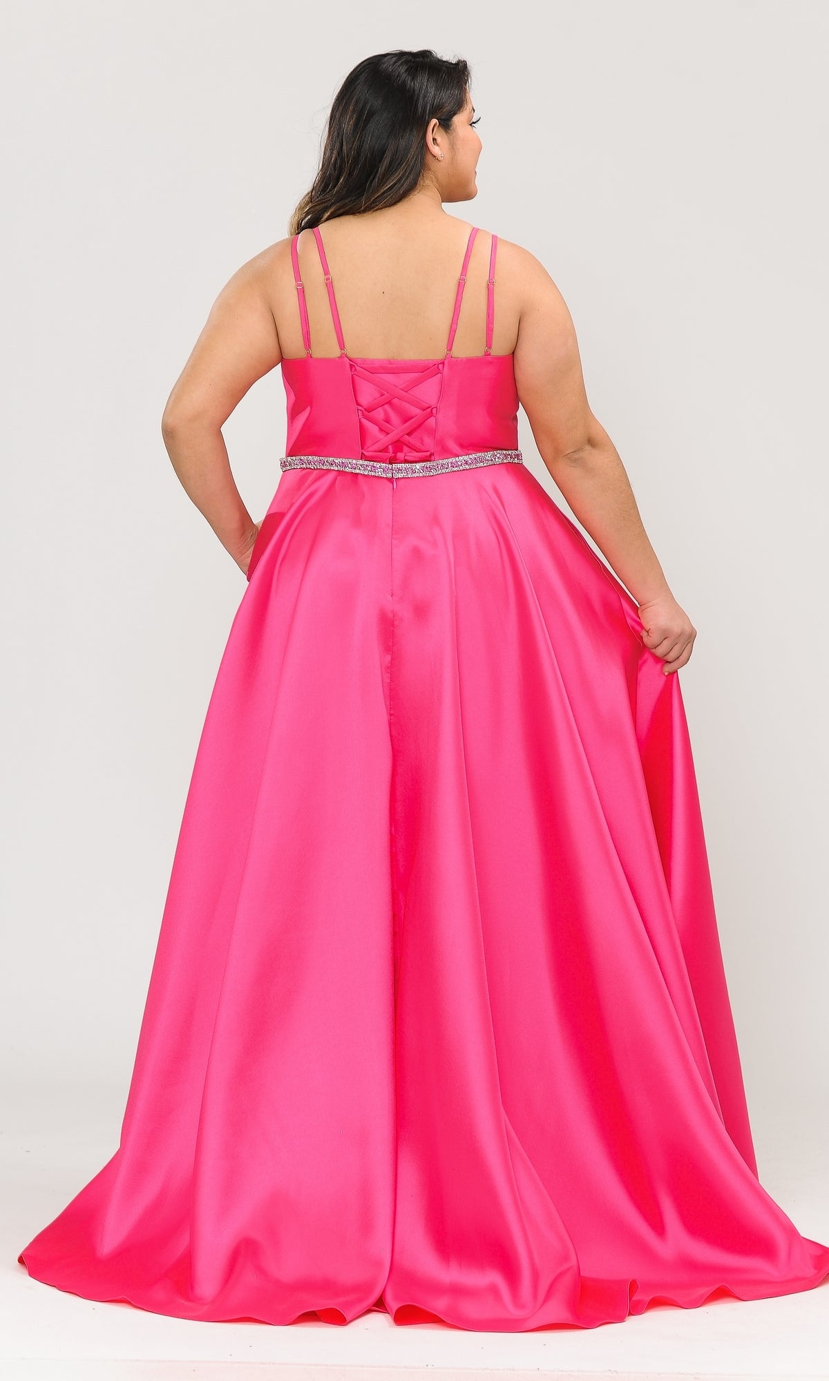 A woman wearing the Plus-Size Long Prom Dress with Pockets W1106 in bright pink satin, featuring double straps and a jeweled waistline, stands facing away while holding the skirt to the side.