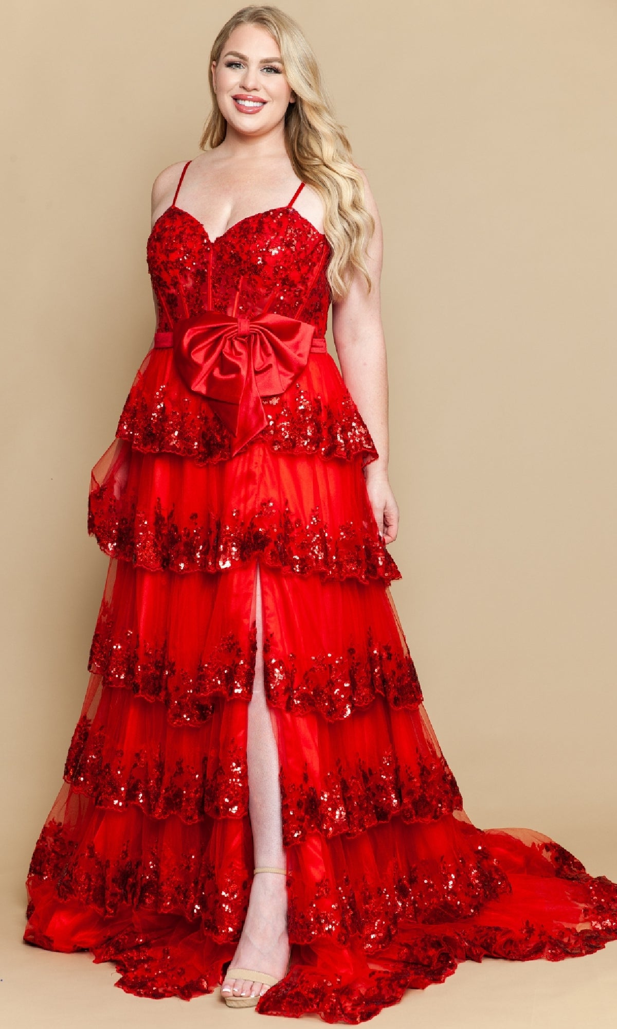 A woman models the Plus-Size Tiered Prom Dress with Bow: PolyUSA W1162, featuring red floor-length ruffled layers, sequin accents, a front slit, and a large waist bow, against a plain beige background.