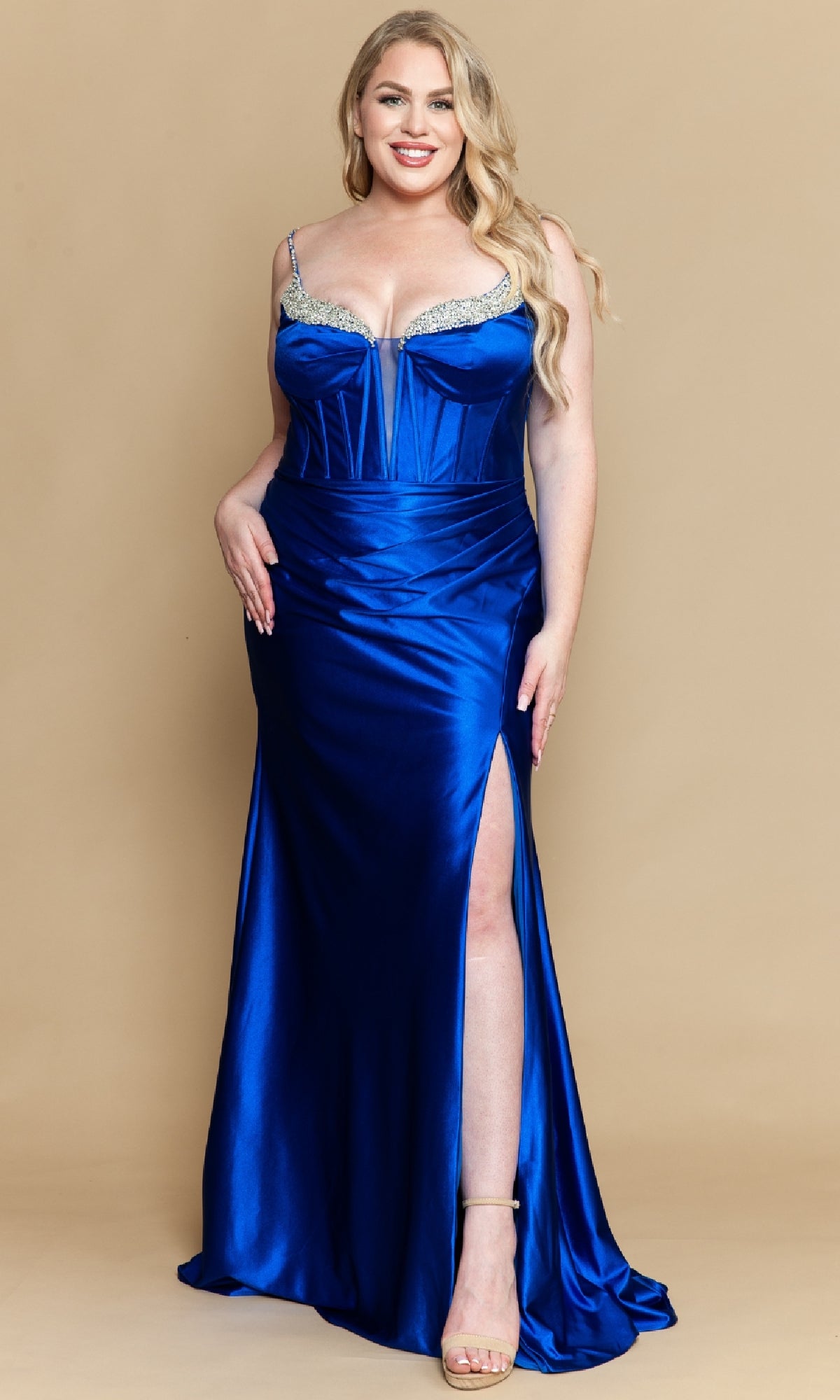 A woman in the Tight Plus-Size Long Satin Prom Dress: PolyUSA W1180, featuring royal blue satin, a thigh-high slit, corset-style bodice, jeweled straps, and nude heels stands against a beige background.
