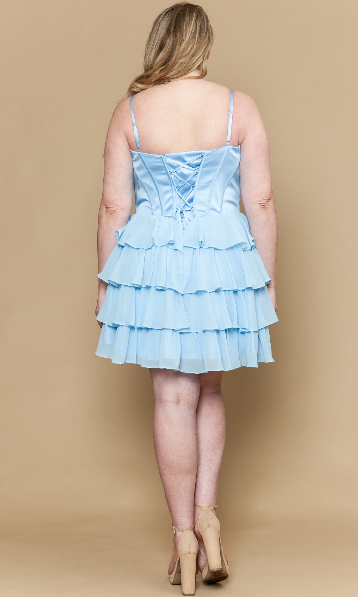 A woman faces away, wearing the Short Plus Size Homecoming Dress: Poly USA W1196—a light blue, ruffled, sleeveless dress with a lace-up back and chiffon a-line skirt—paired with beige heels against a beige background.