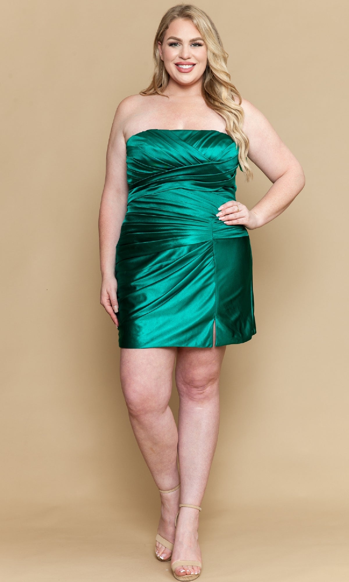 A woman wearing the Plus-Size Strapless Party Dress: Poly USA W1206 in emerald green satin, featuring a short length and side slit, stands against a beige background, smiling with one hand on her hip.