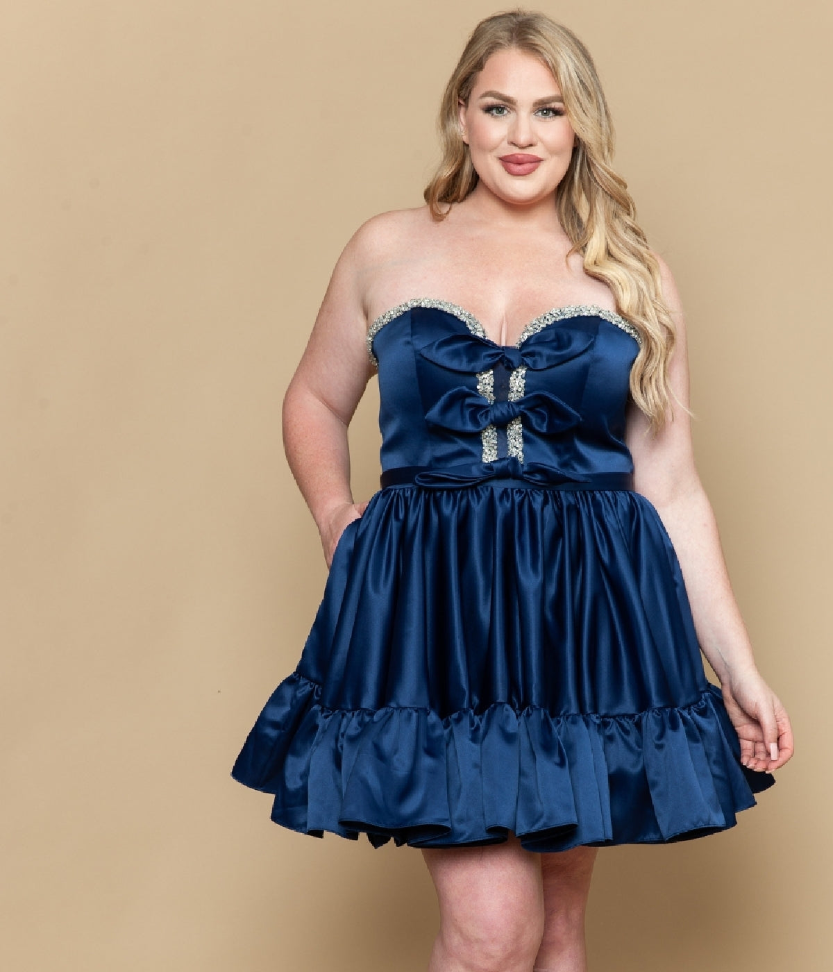 A woman wearing the Short Plus Size Homecoming Dress: Poly USA W1212 poses against a beige background, smiling confidently. The strapless satin fit-and-flare style with a ruffled skirt highlights her figure elegantly.