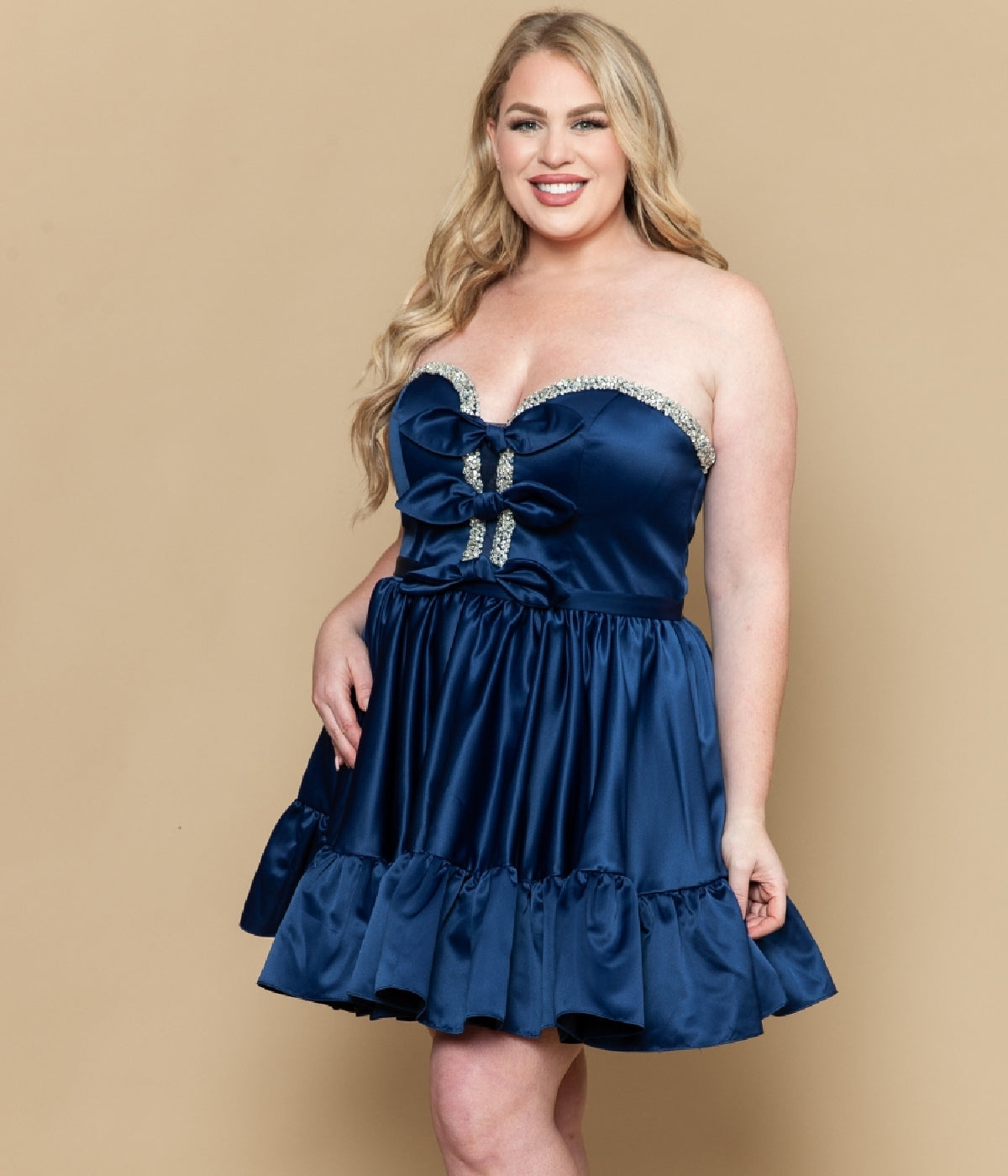 A woman smiles in the Poly USA W1212 Short Plus Size Homecoming Dress, a strapless satin fit-and-flare style with ruffle hem and rhinestone accents, standing against a plain beige background.