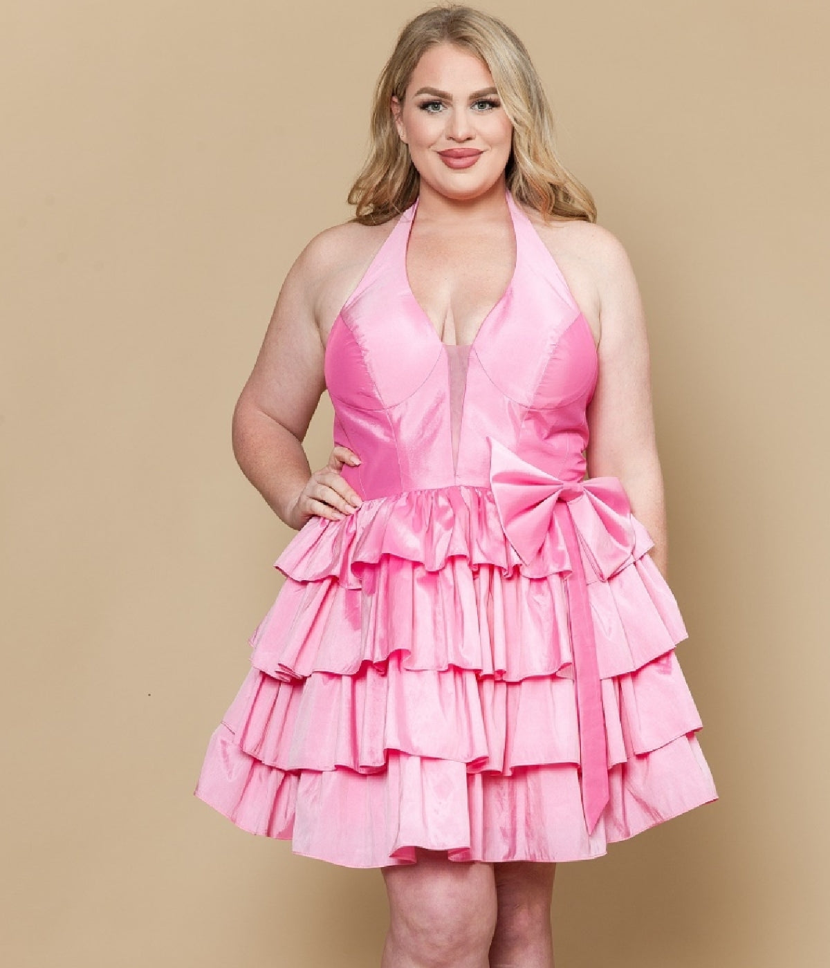 A woman poses against a beige background in the Plus-Size Short Halter Party Dress: Poly USA W1216 in pink, featuring tiered ruffles and a large bow on the skirt.