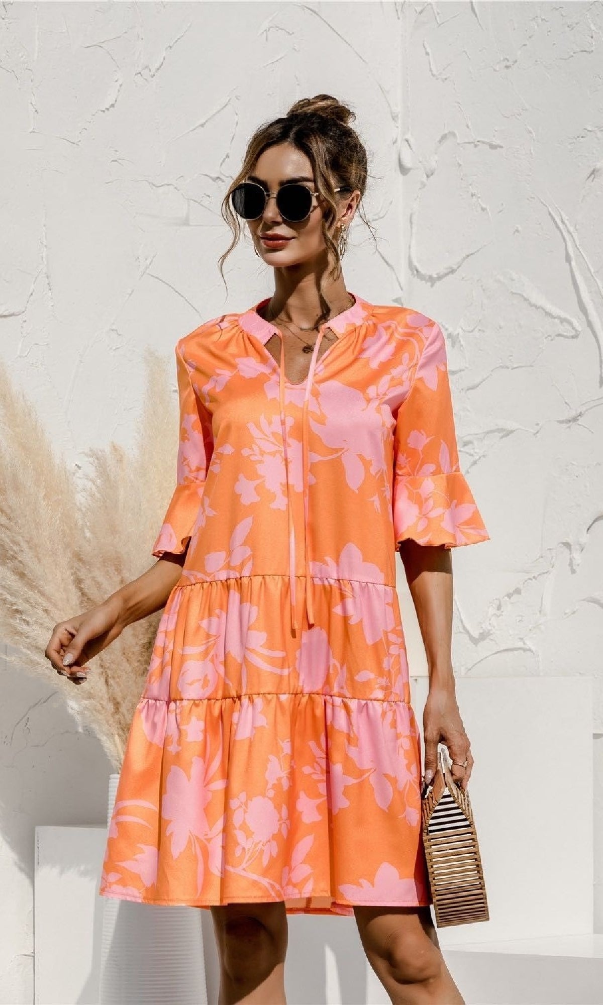 A woman in a bright orange and pink Tiered A-Line Short Sleeve Casual Print Dress with ruffled sleeves, sunglasses, and a woven handbag stands against a textured white wall.
