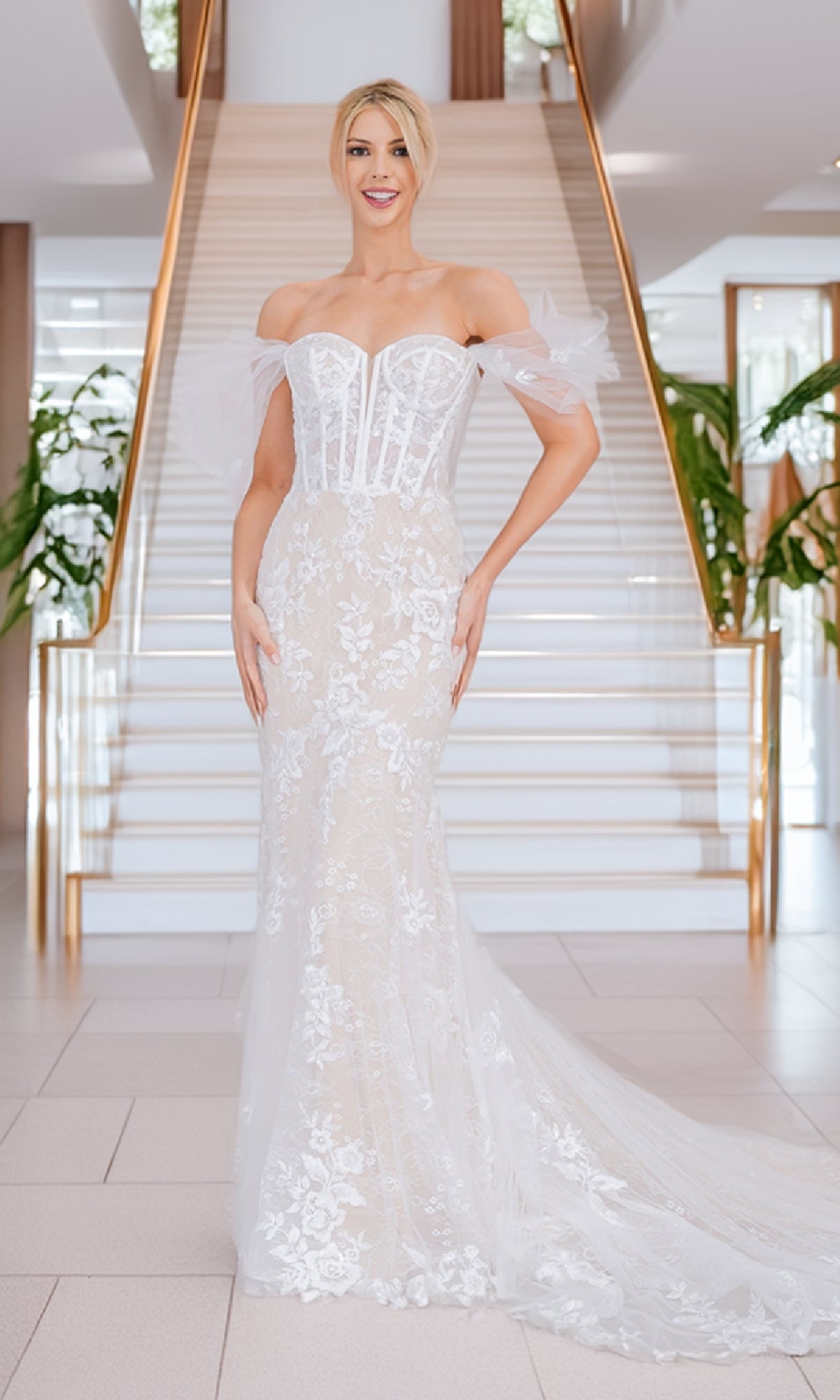 A woman in the Amelia Couture WL0100 Long White Bridal Gown stands before a modern staircase, framed by lush plants on each side.