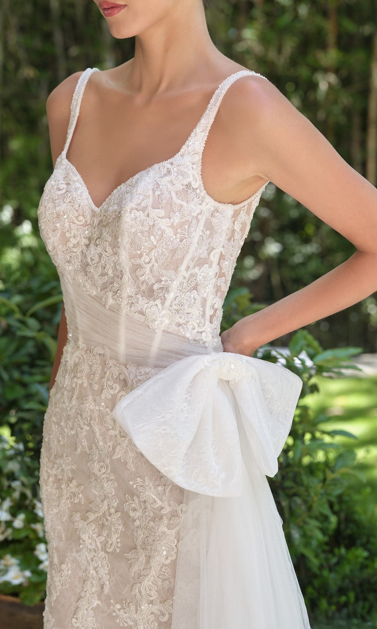 A woman in the Andrea and Leo WL068 long wedding dress—white lace with a sheer corset bodice, beaded straps, and a large tulle bow at the hip—stands outdoors amid lush greenery.