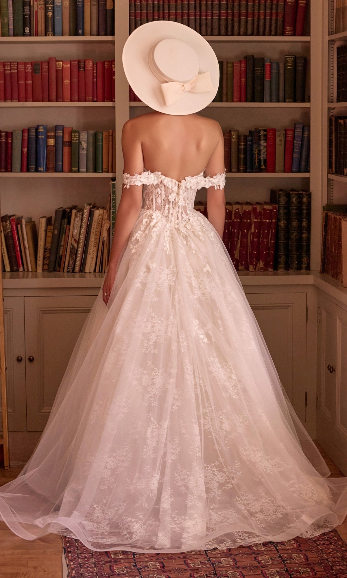 A person wearing an off-the-shoulder Long French White Wedding Dress: Andrea & Leo WL093 and a wide-brimmed hat, standing with their back to the camera in front of a bookshelf.