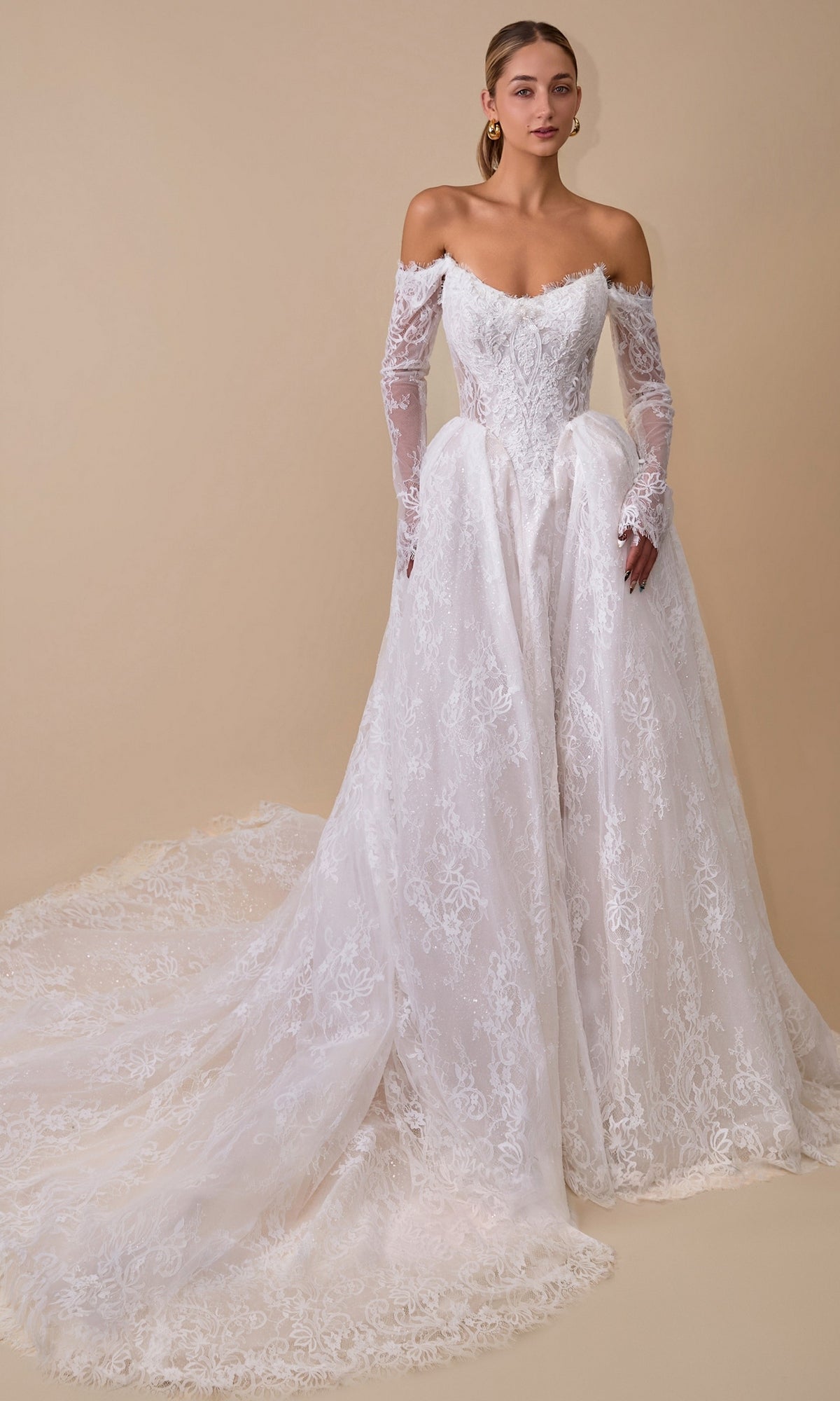 An elegant woman models the Andrea & Leo WL079 long wedding dress in off-white, showcasing its off-the-shoulder neckline, long lace sleeves, and sweeping train against a plain beige background.