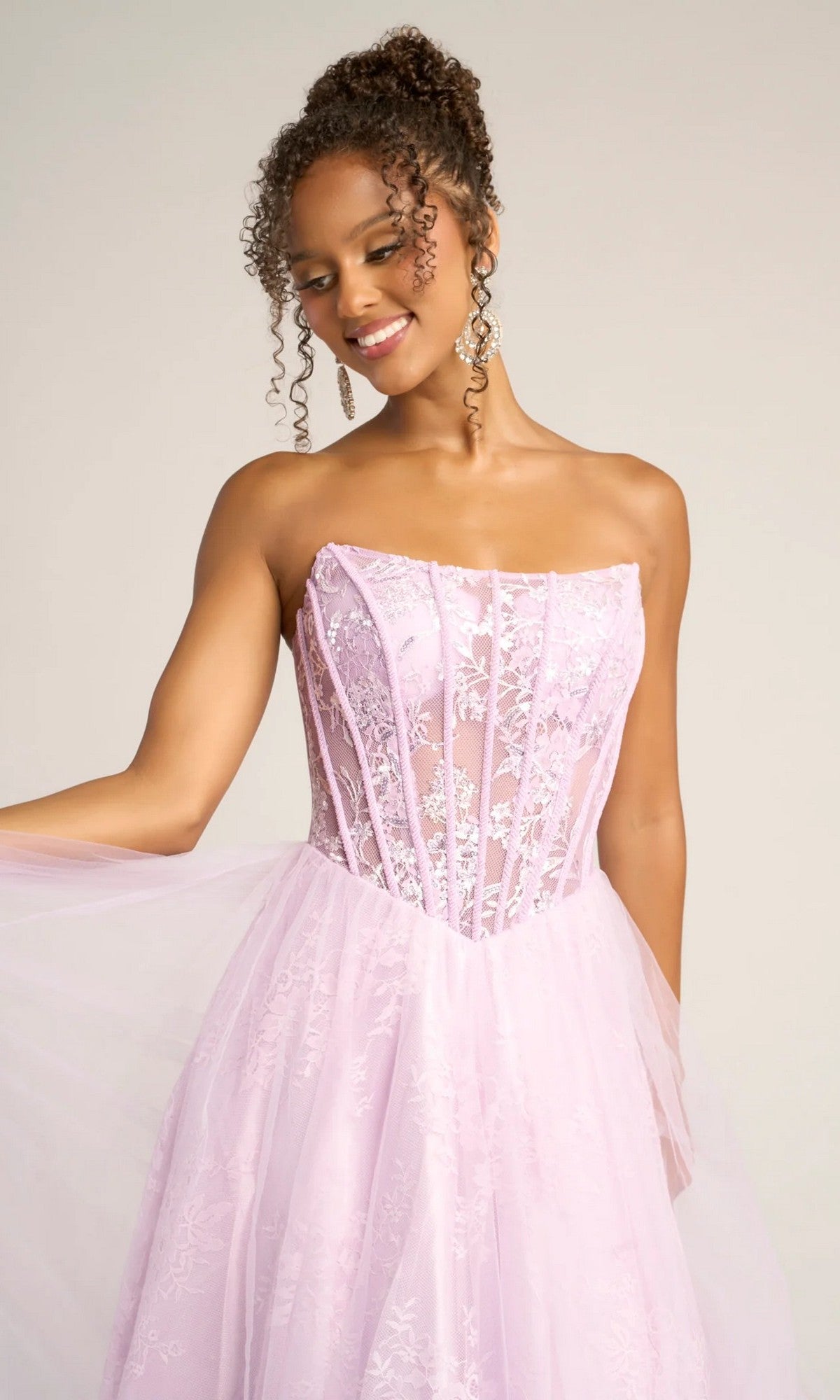 A woman in the Portia and Scarlett XO023 long formal dress with a strapless lace ball gown silhouette, sheer corset bodice, and tulle and lace skirt holds part of the skirt while smiling.
