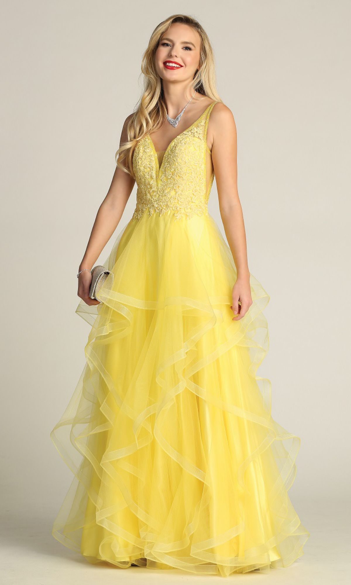 A woman in the Sheer-Sides Long Tiered Prom Dress (Chicas XR6006), featuring a yellow ruffled skirt and beaded v-neck bodice, smiles at the camera while holding a small clutch.