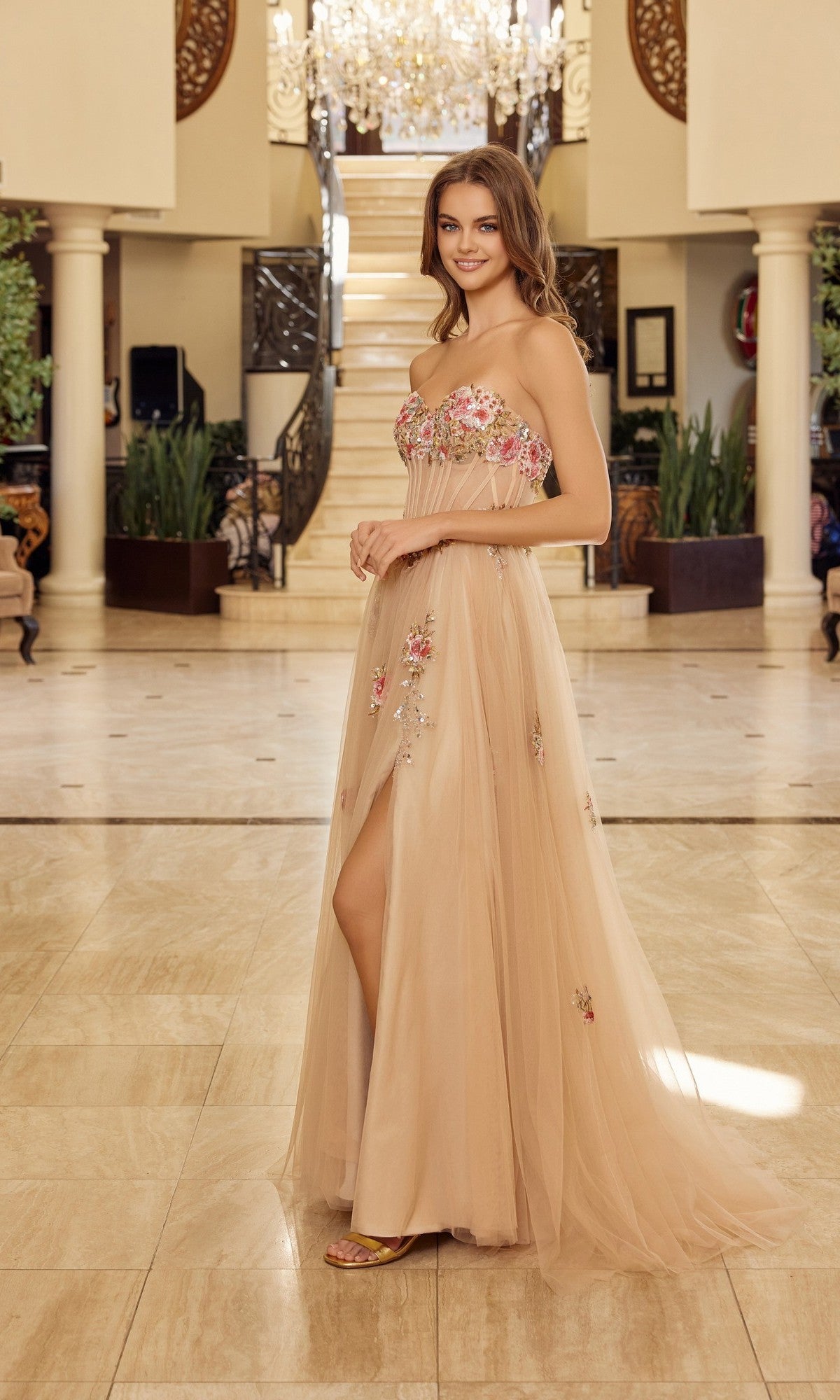 A woman wearing the Nox Anabel Y1803 long prom dress with delicate floral embroidery stands indoors on a marble floor, with a staircase and chandelier in the background.