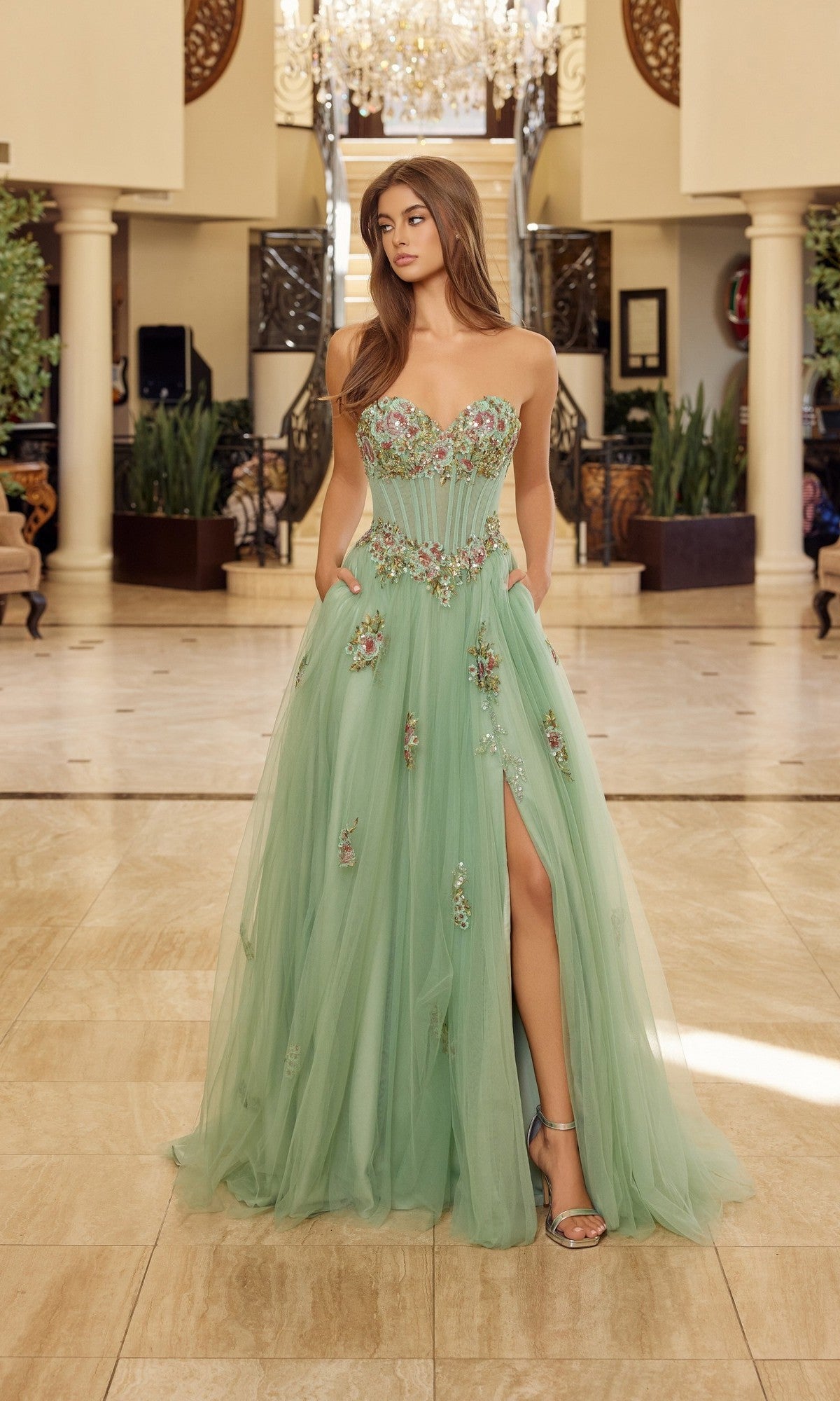 A woman stands indoors in the Nox Anabel Y1803 long prom dress in sage green, featuring floral embroidery, a strapless bodice, thigh-high slit, and floor-length tulle for an elegant touch.