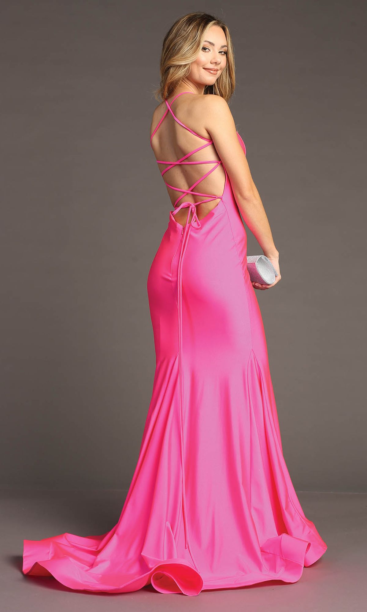 Wearing the affordable Strappy Open-Back Long Prom Dress: Chicas YG5024—a floor-length, bright pink gown with crisscross straps—a woman holds a silver clutch against a plain background.