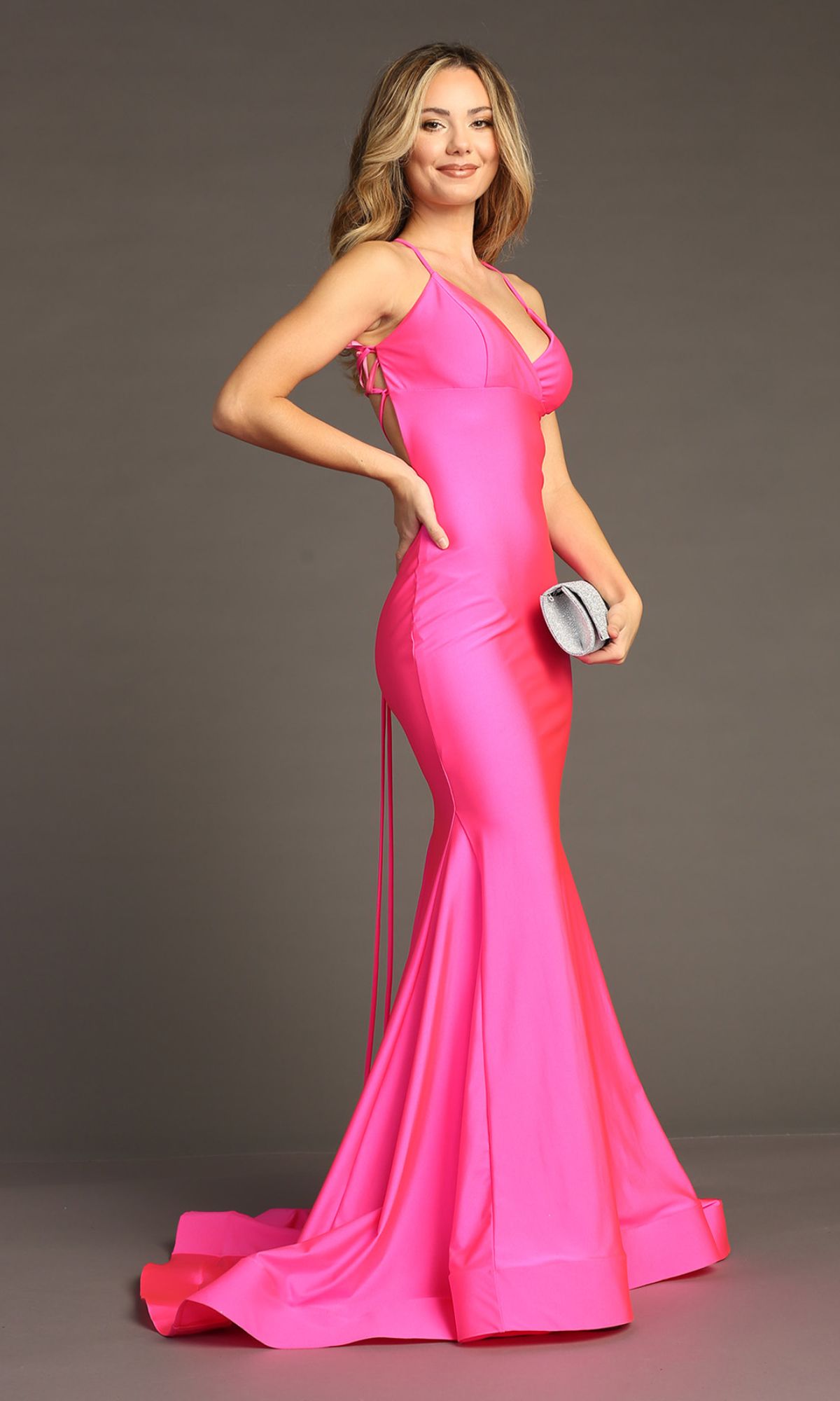 A woman in the Strappy Open-Back Long Prom Dress Chicas YG5024, a bright pink, sleeveless, floor-length mermaid gown, holds a silver clutch and stands against a plain background.