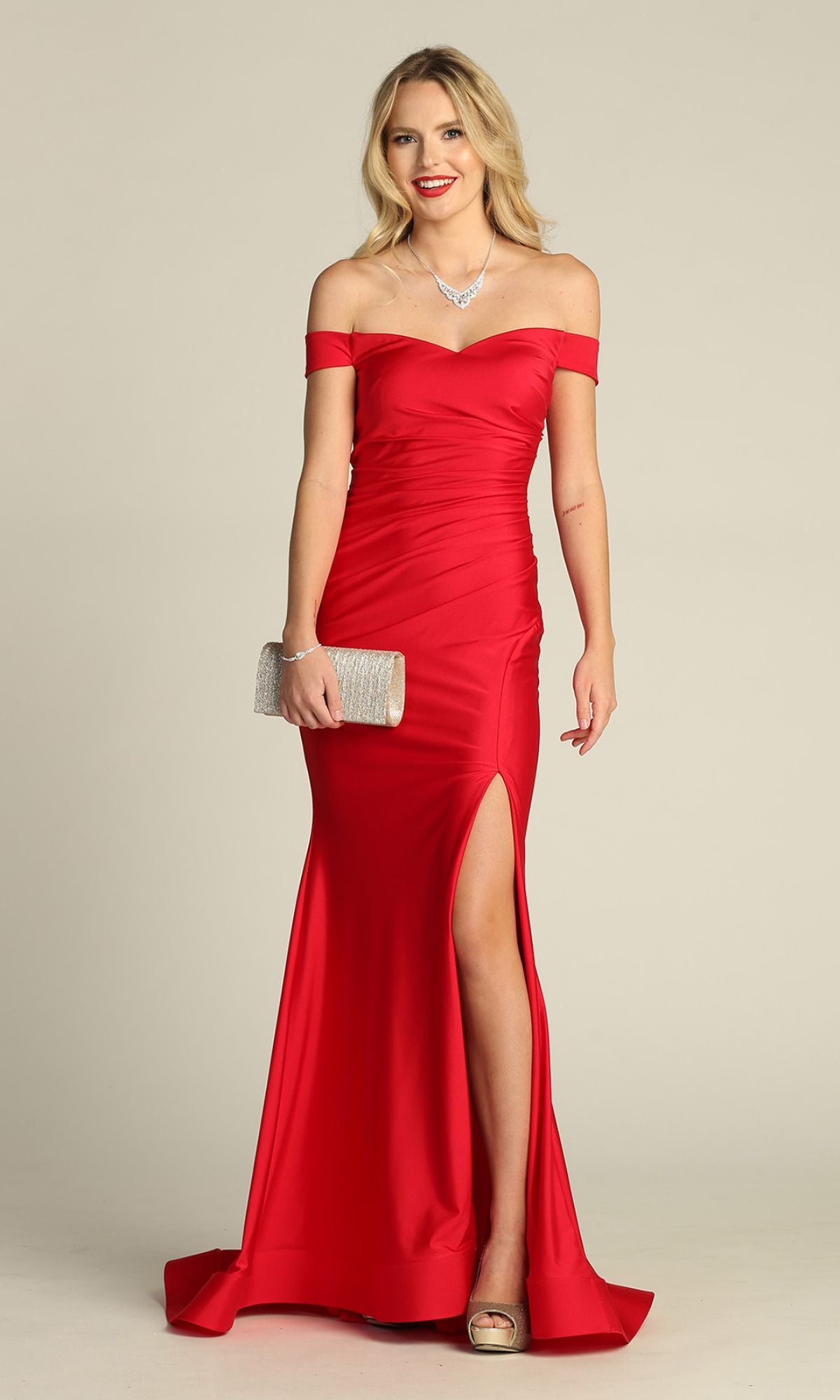 A woman in the Off-the-Shoulder Long Prom Dress: Chicas YG5025—a red, floor-length gown with a thigh-high slit—holds a silver clutch and stands before a plain backdrop.