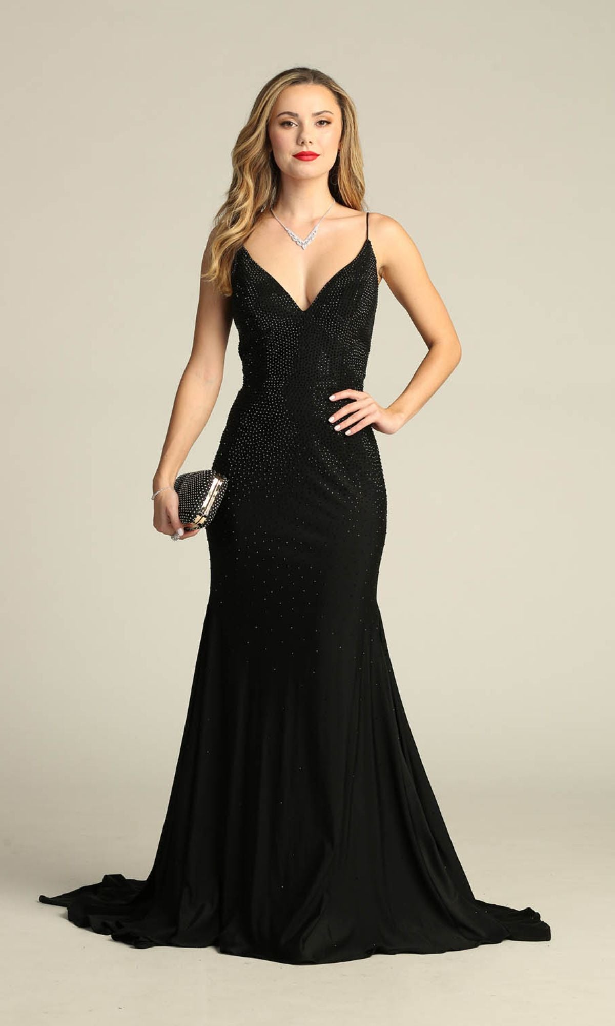 A woman stands in the Lace-Up Long Beaded Prom Dress: Chicas YG5026, a fitted black formal gown with thin straps, holding a clutch and wearing a necklace.