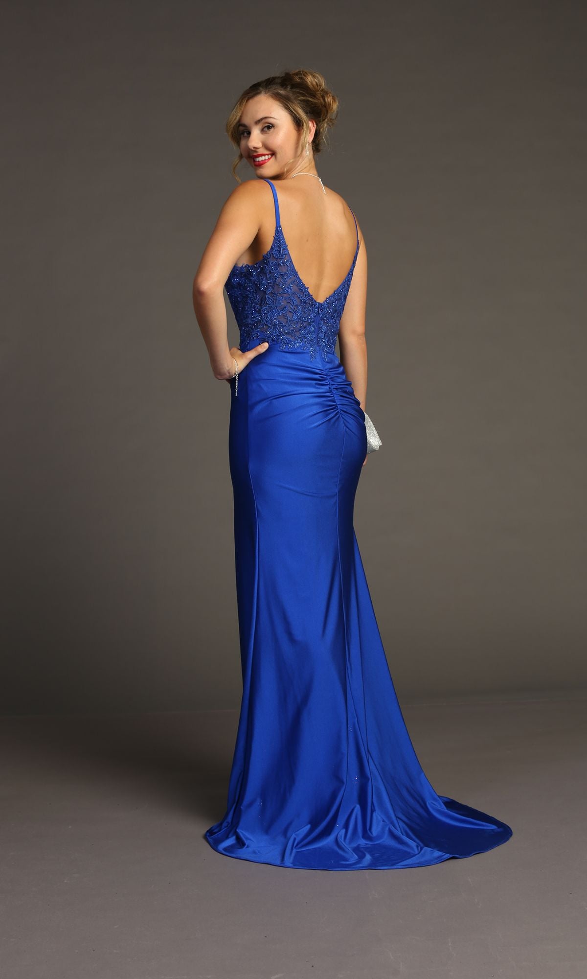 A woman in the Lace-Bodice Slinky Long Prom Dress Chicas YG5033, a sleeveless royal blue gown with a low back and ruched detailing, stands facing away and looks over her shoulder.