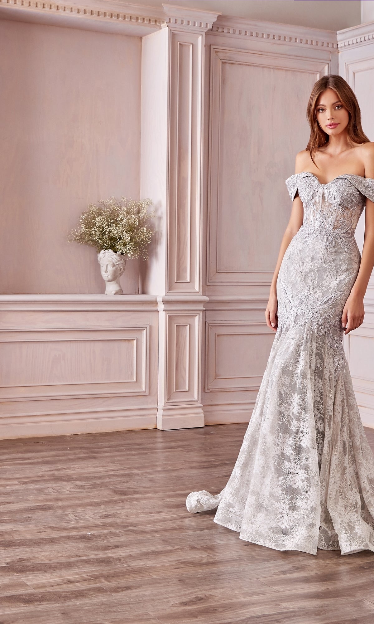 A woman in a fitted Off-the-Shoulder Long White Lace Prom Gown stands in an elegant room with light wood paneling and a bust vase filled with flowers on a shelf behind her.