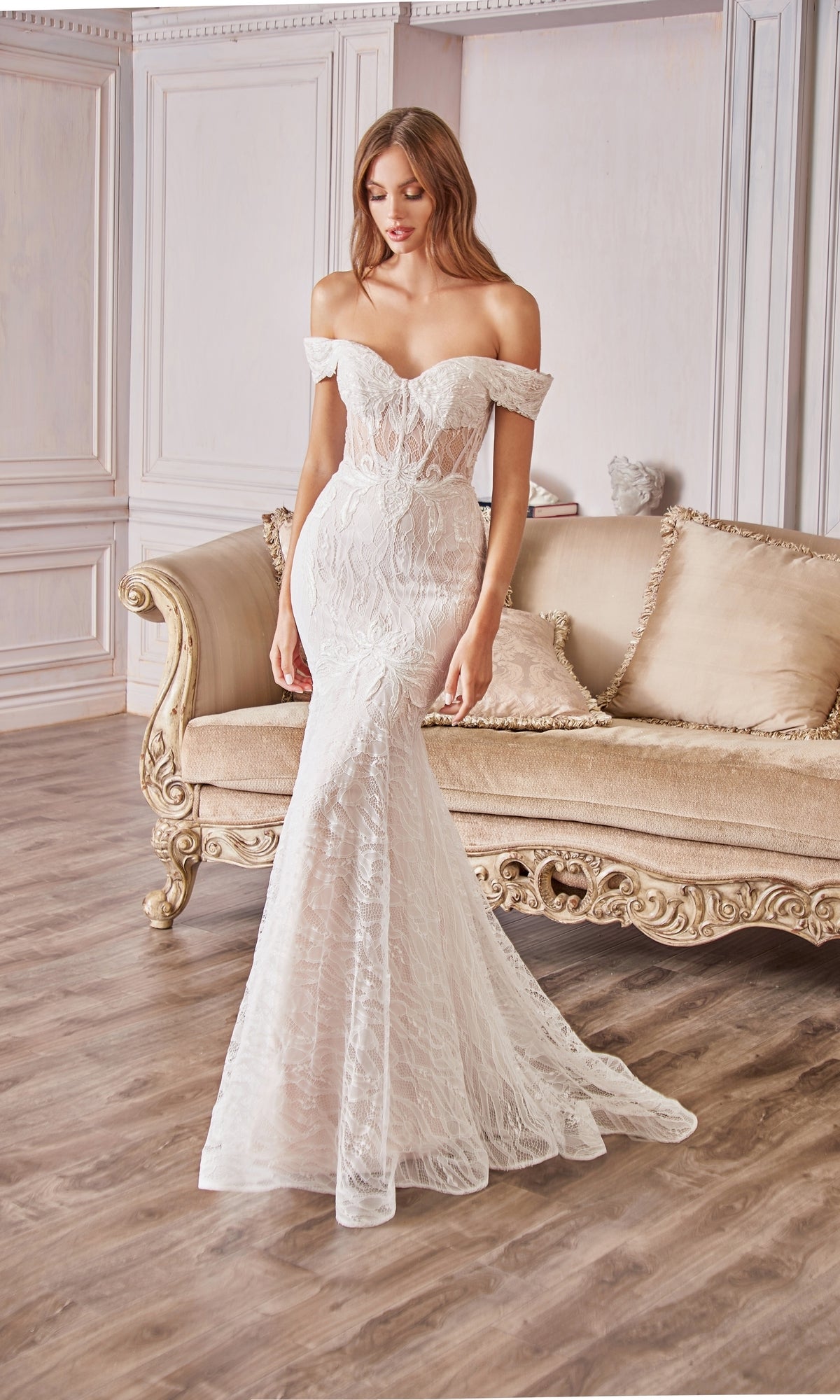A woman in an Off-the-Shoulder Long White Lace Prom Gown stands by a beige ornate sofa in an elegant room with wood floors, her fitted dress radiating timeless sophistication.