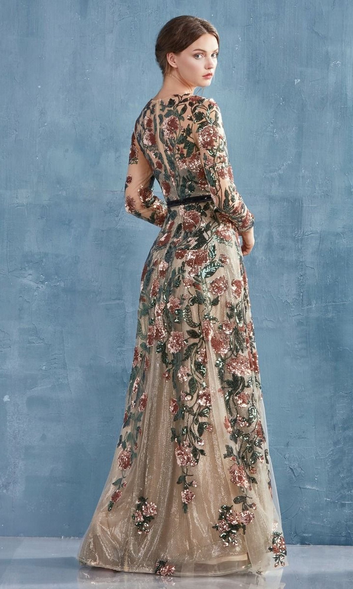 A woman stands against a textured blue wall, wearing the Long Sleeve Sequin-Floral Long Formal Dress A0926—an elegant gown with intricate green, pink, and gold floral embroidery.