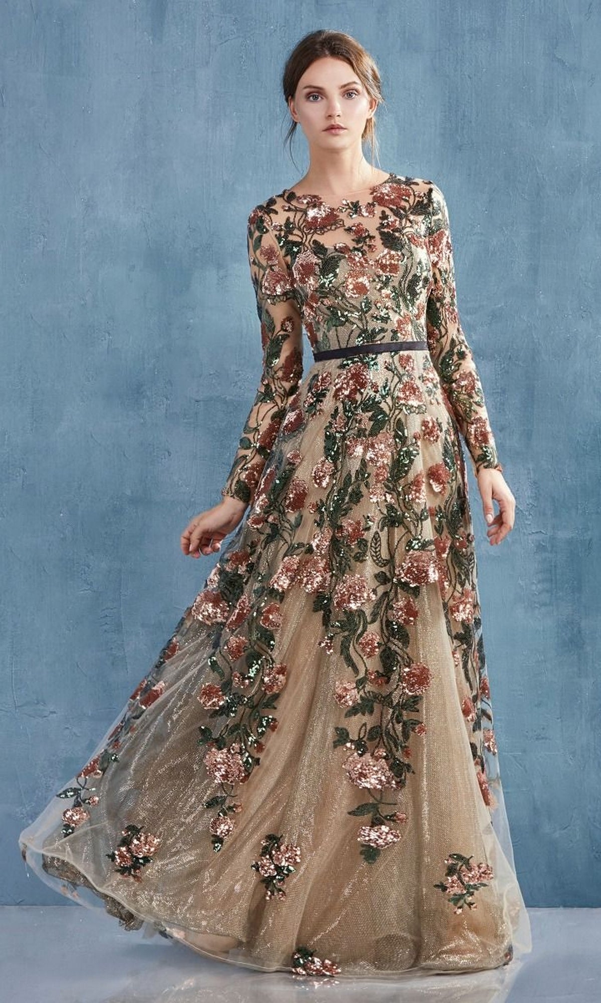 A woman stands against a blue textured wall, wearing the Long Sleeve Sequin-Floral Long Formal Dress A0926—an elegant gown with sequins, floral accents, sheer fabric, and delicate embroidery.