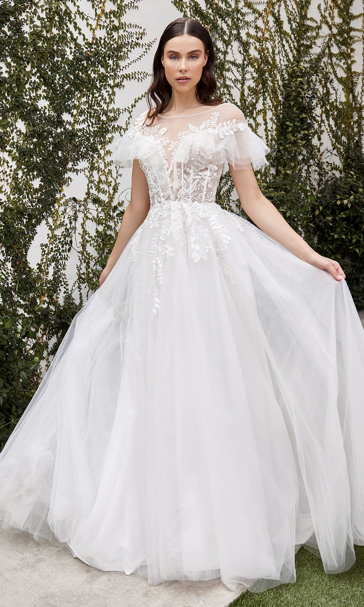 A woman stands outdoors near lush greenery wearing the Ladivine White A-Line Wedding Dress A1070W, featuring off-the-shoulder ruffled floral lace details.