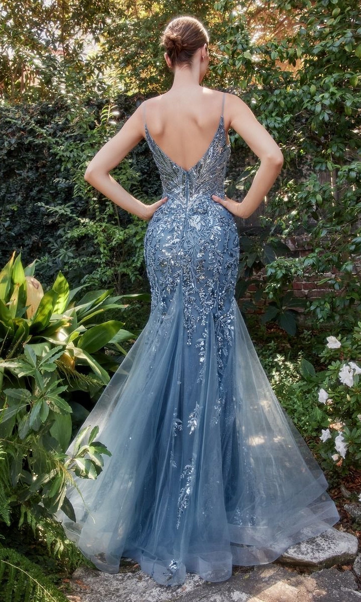A woman stands outdoors facing away, wearing the Sequin-Print V-Neck Long Mermaid Prom Dress A1118 with a deep V-back and silver details, surrounded by lush green plants.