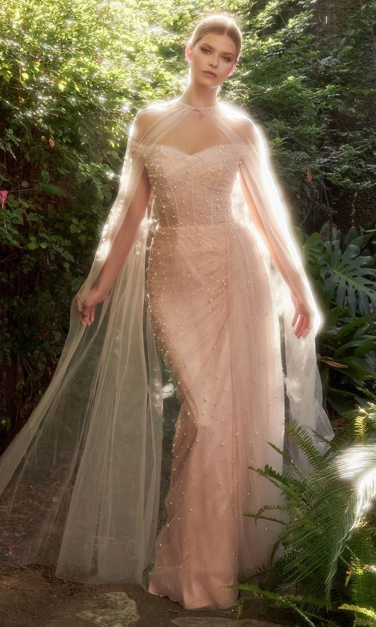 A woman stands outdoors in a Long Pink Pearlized Formal Dress with Cape, surrounded by greenery and soft sunlight.