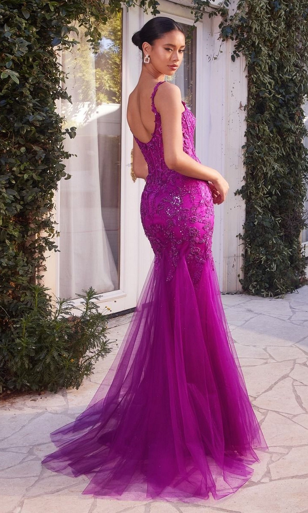 A woman stands outdoors near a white door framed by greenery, wearing the fitted Long Lace Mermaid Prom Dress Andrea and Leo A1231 in magenta, featuring a sheer bodice with embroidered lace and sequin details.