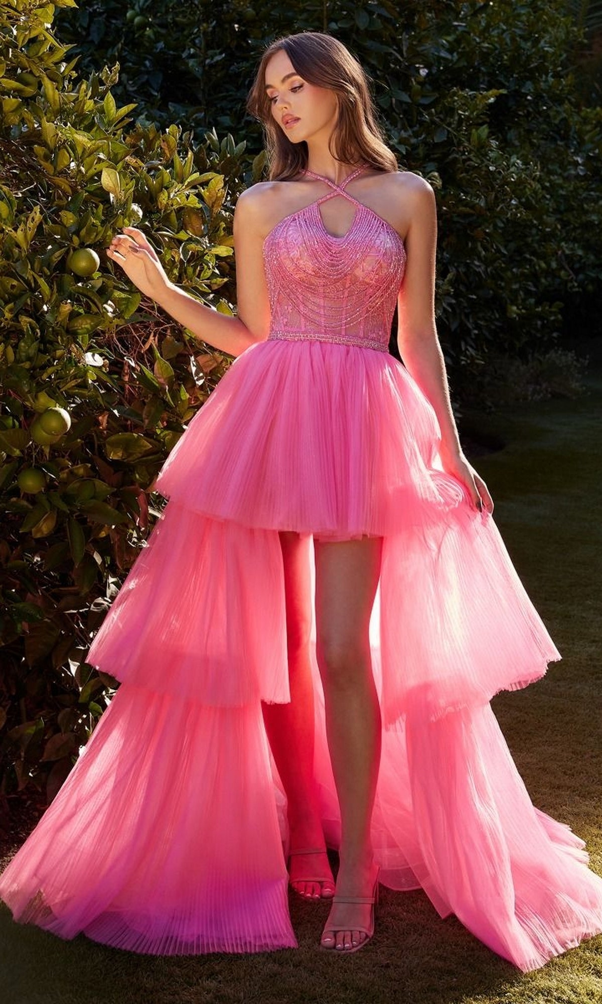 A woman stands outdoors in the High-Low Beaded-Bodice Ruffle Prom Ball Gown A1239, featuring a bright pink color, fitted beaded bodice, and pleated high-low layers, surrounded by green foliage.