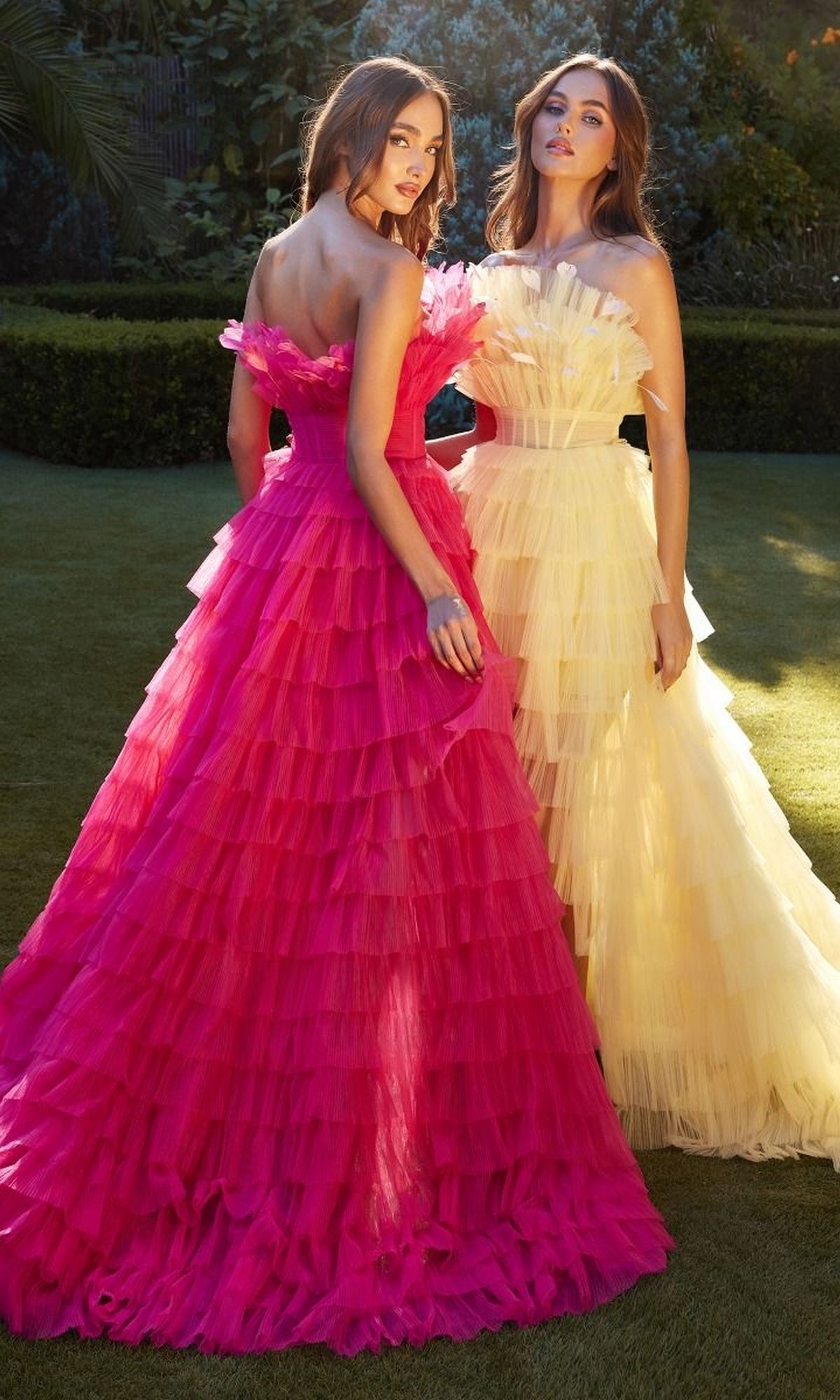 Two women stand on grass in Andrea and Leo's Ruffled High-Low Ball Gown A1240—one gown in bright pink, the other in pale yellow. Both feature tiered ruffles and dramatic feather trim, as they pose elegantly against lush greenery.