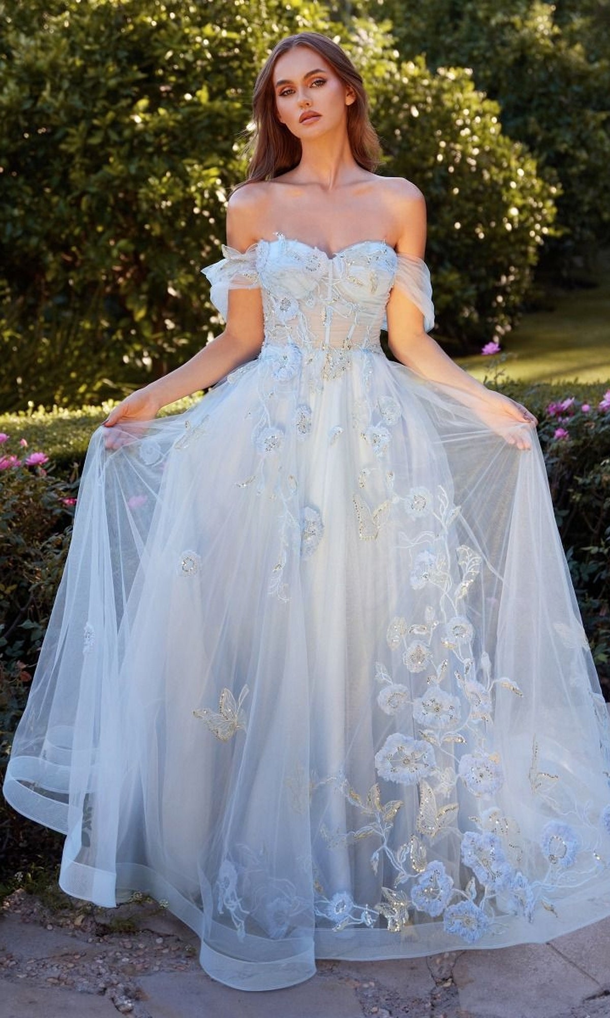 A woman poses outside in the Andrea and Leo A1246 Long Blue Prom Ball Gown, an off-the-shoulder evening dress with floral embroidery and sheer fabric, set against a lush backdrop of greenery and flowers.