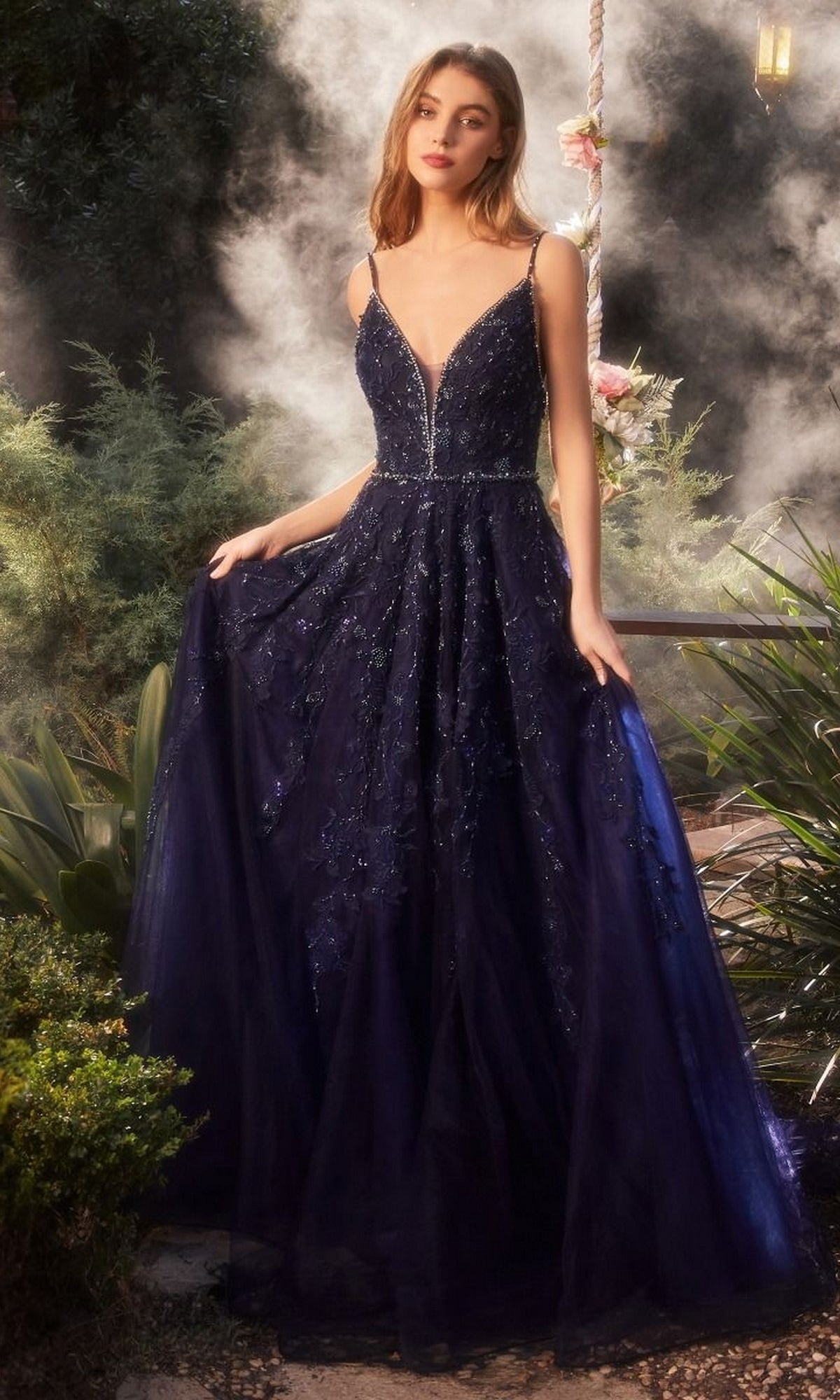 A woman stands outdoors in the Long Formal Dress A1251 by Andrea and Leo, a navy ball gown with a fitted bodice and floor-length tulle skirt, shimmering amid lush greenery.