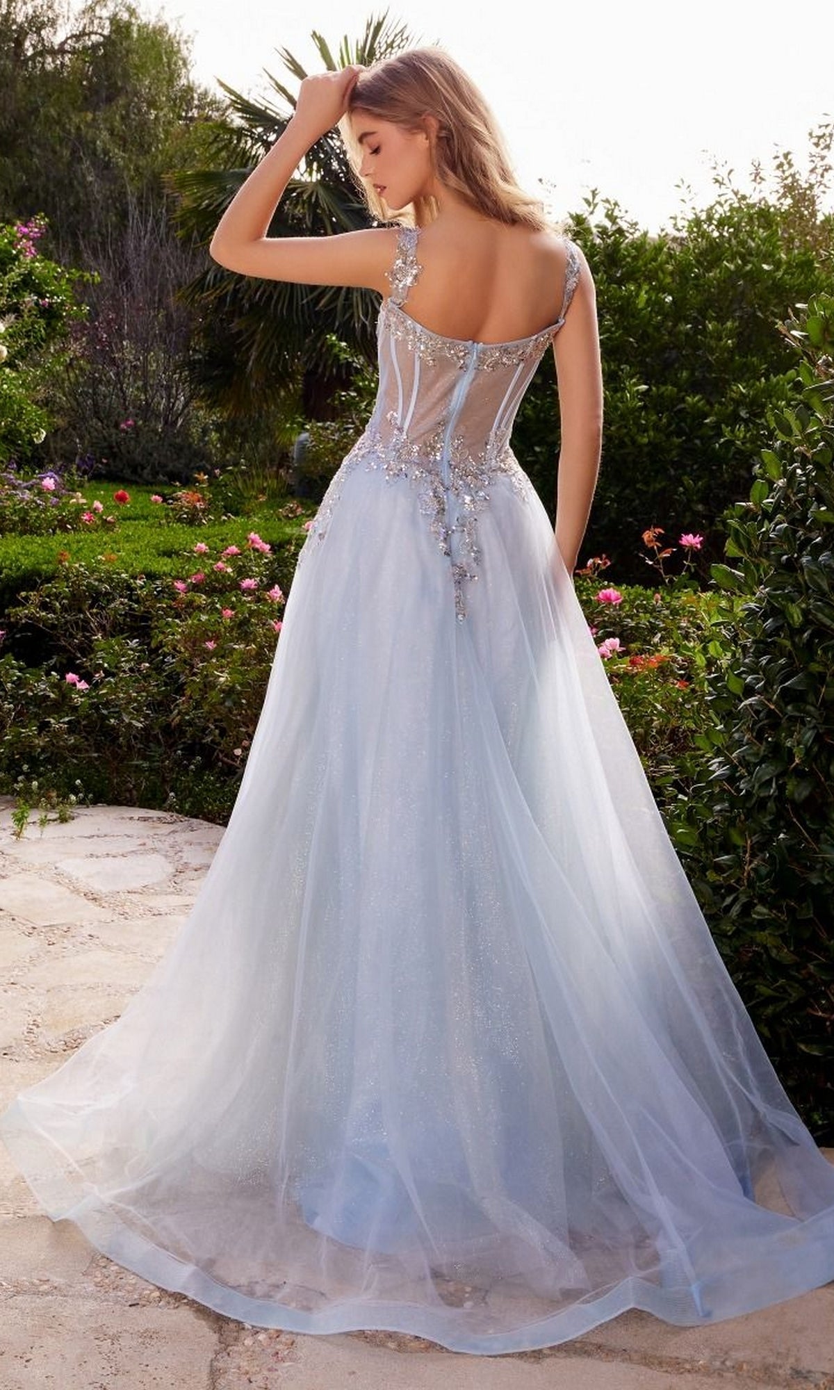 Outdoors in a garden, a woman wears the Long Pastel A-Line Prom Dress: Andrea & Leo 1258—a light blue, floor-length gown with delicate floral embroidery, sheer fabric, an open back, and elegant embellishments.
