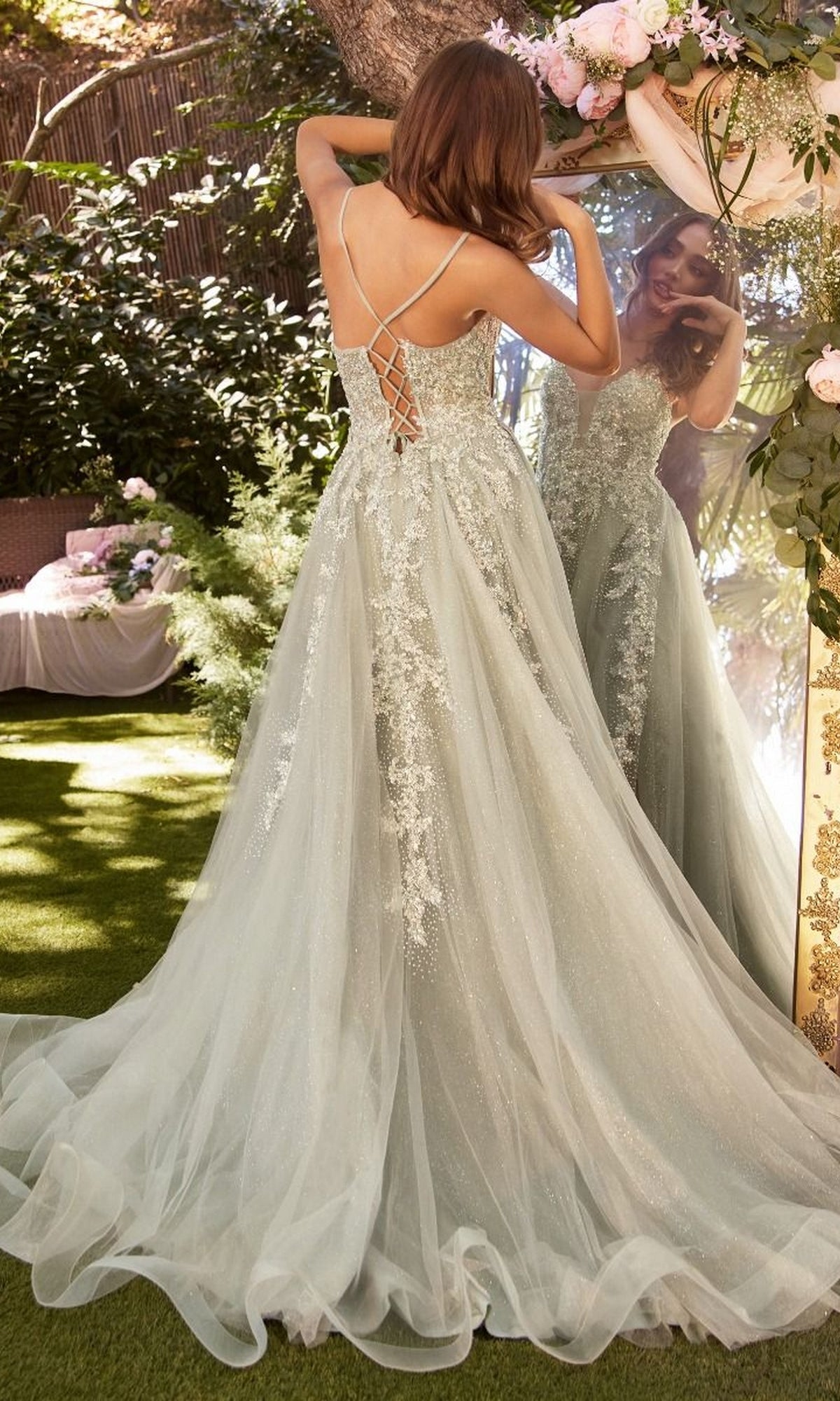 A woman in the Sage Green Long Glitter-Tulle Prom Dress A1261 stands outdoors, facing a mirror and adjusting her hair.