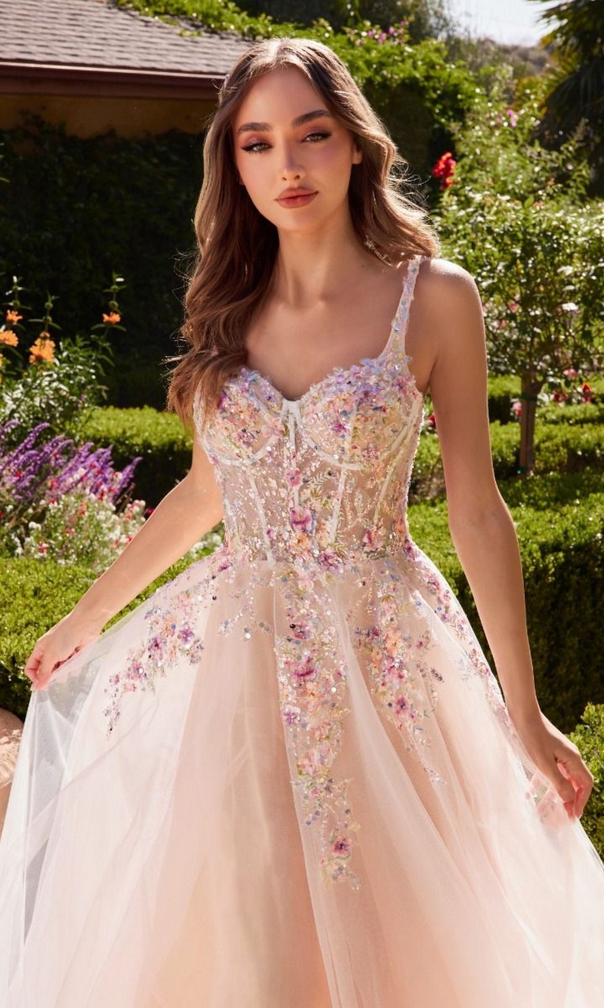 A woman in the Floral-Bodice Off White Long Prom Ball Gown A1288 stands outdoors amidst lush greenery and vibrant flowers.