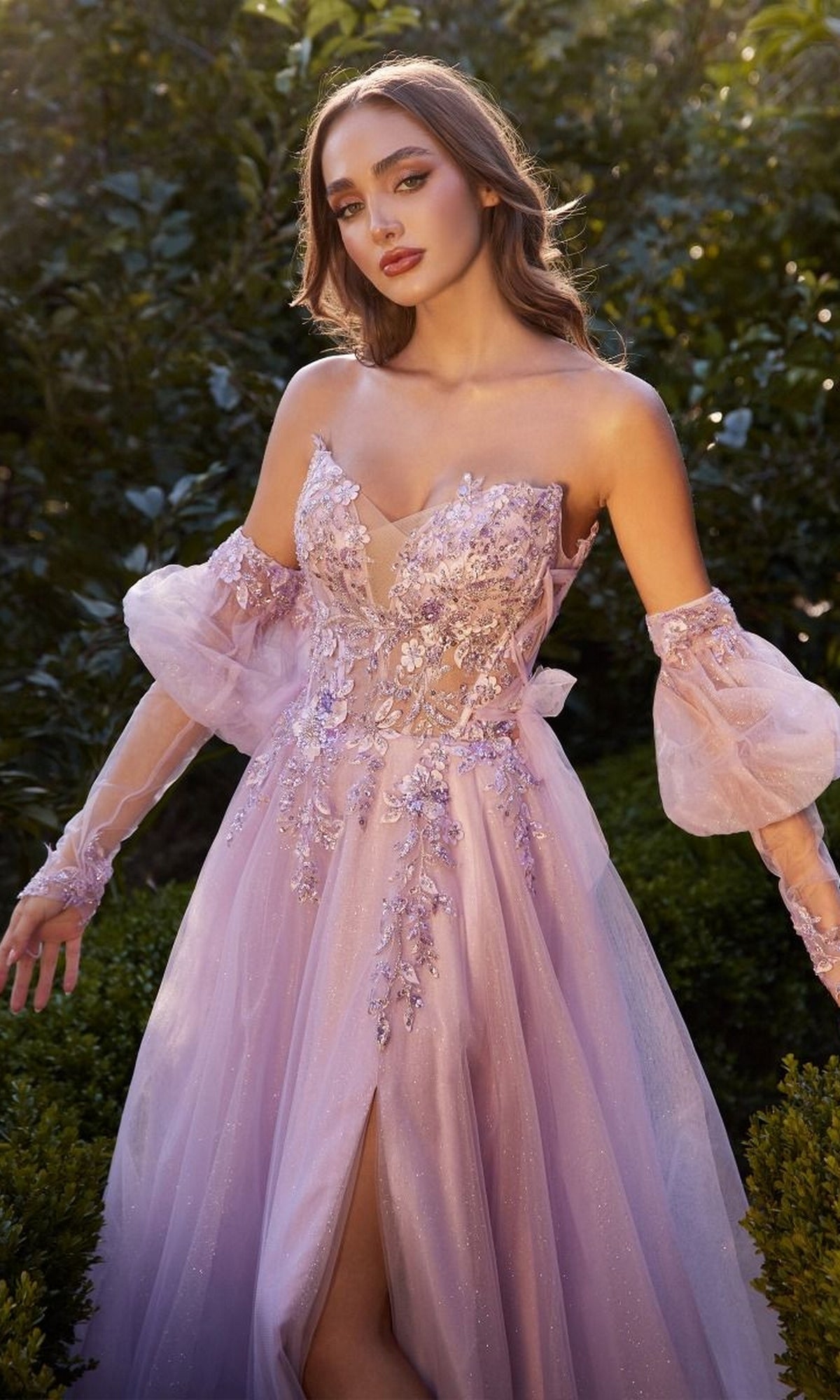 A woman stands outdoors in the Gloved Strapless Prom Gown: Andrea and Leo A1303, a light purple off-the-shoulder dress with floral embroidery and sheer puff sleeves, shimmering amidst lush greenery.
