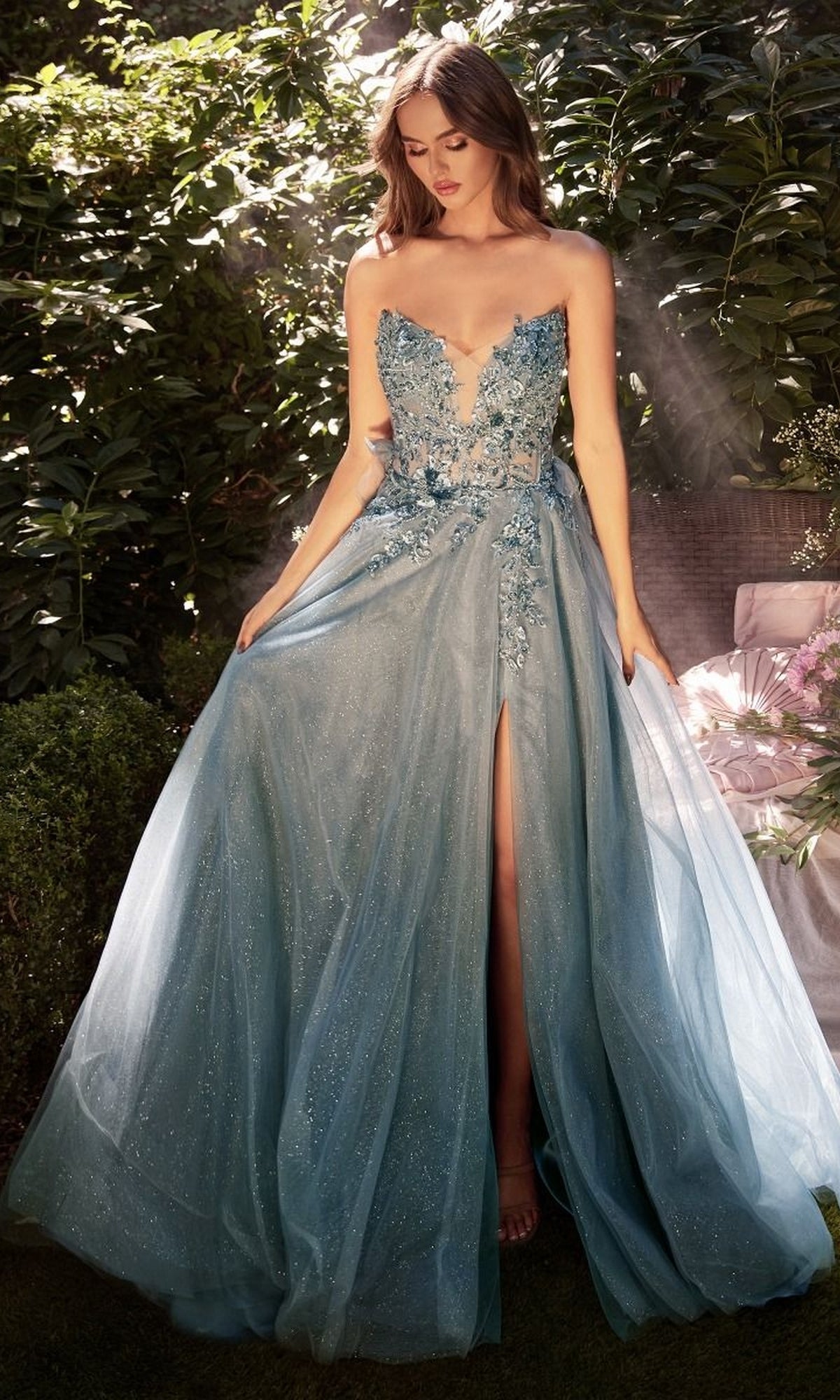 Basking in sunlight, she wears the Andrea and Leo A1303 Gloved Strapless Prom Gown—an elegant blue dress with floral appliqués, a deep neckline, high slit, and sparkling details that shimmer beautifully outdoors.